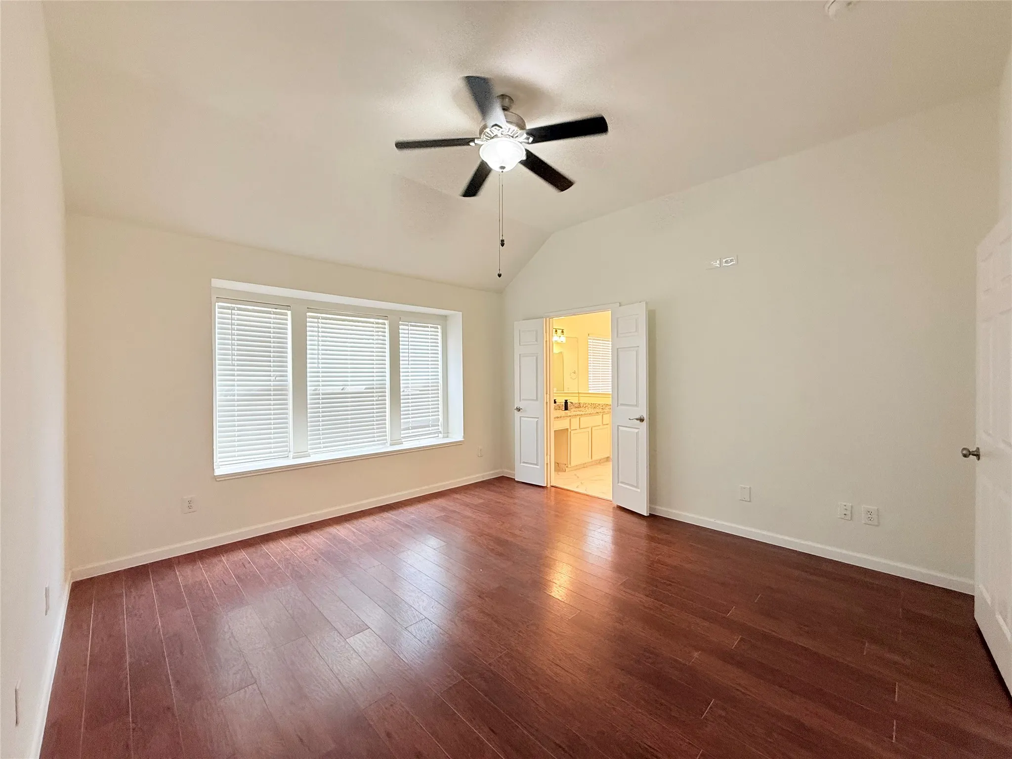 Vaulted ceiling with a ceiling fan, recessed window alcove with blinds, wood-finish flooring, and double doors opening to a bathroom