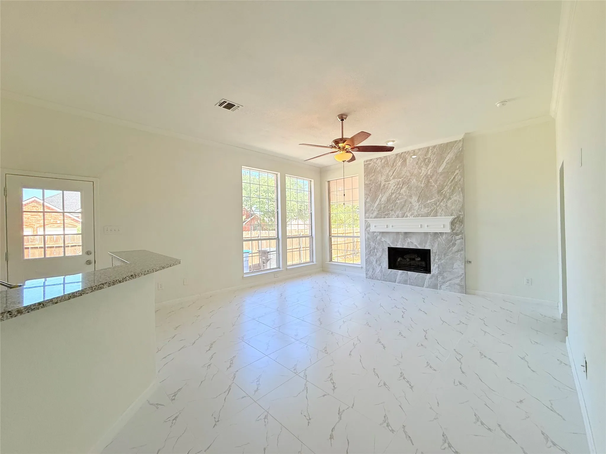 Spacious living area featuring large windows, white tile flooring, a built-in fireplace with a stone surround, and a ceiling fan