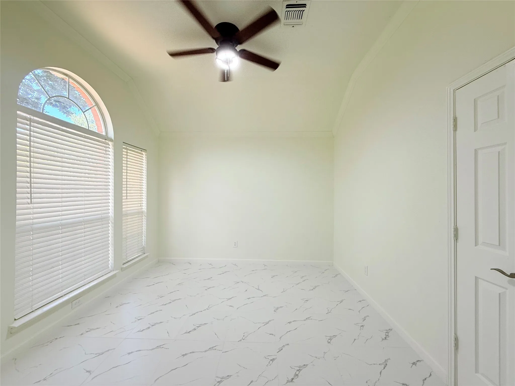 Vaulted ceiling with crown molding, ceiling fan light fixture, arched window, white blinds, and white marble-look flooring