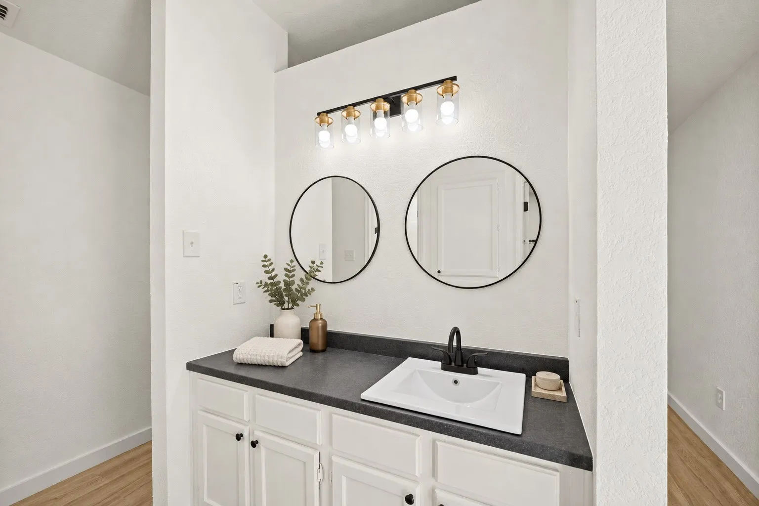 White vanity with panel cabinetry and black hardware, dark countertop, single basin sink with matte black faucet, and two round mirrors