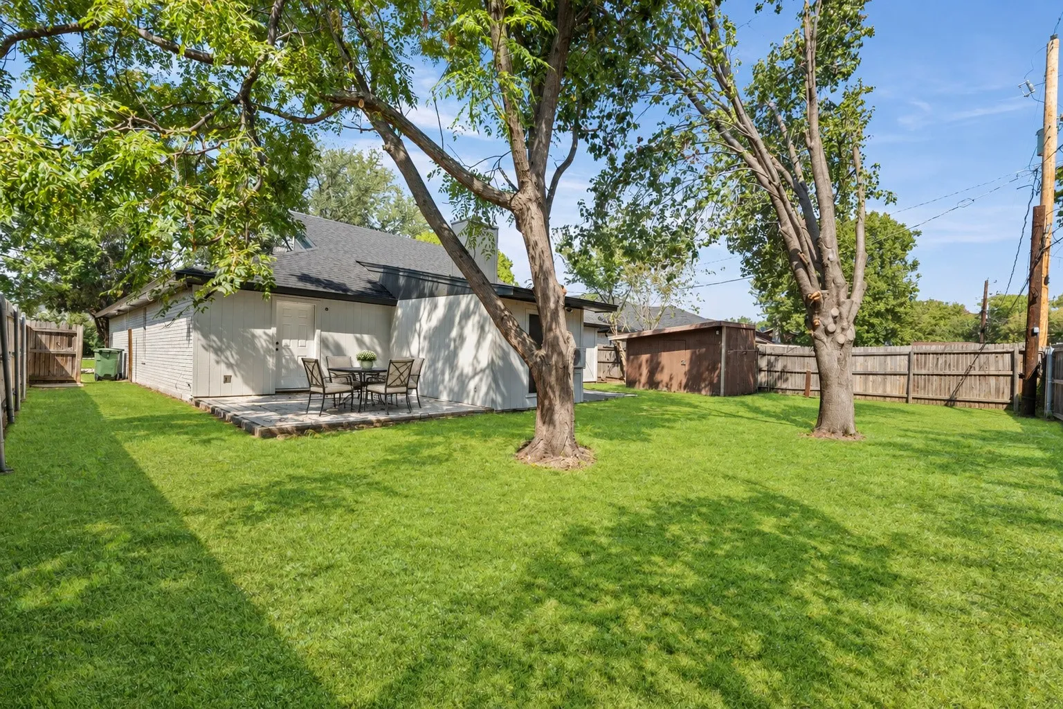Expansive green lawn with mature trees, stone patio, wood privacy fencing, and a detached storage shed
