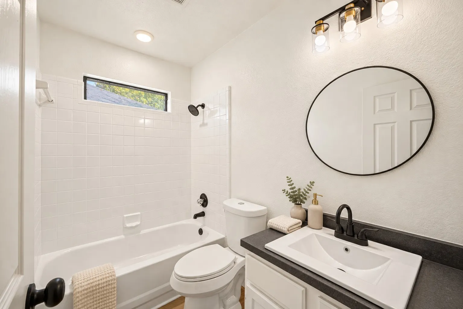 Bathroom featuring a white subway tile shower surround, a bathtub with matte black fixtures, a white vanity with a dark countertop, a rectangular undermount sink, and a circular mirror with a black frame