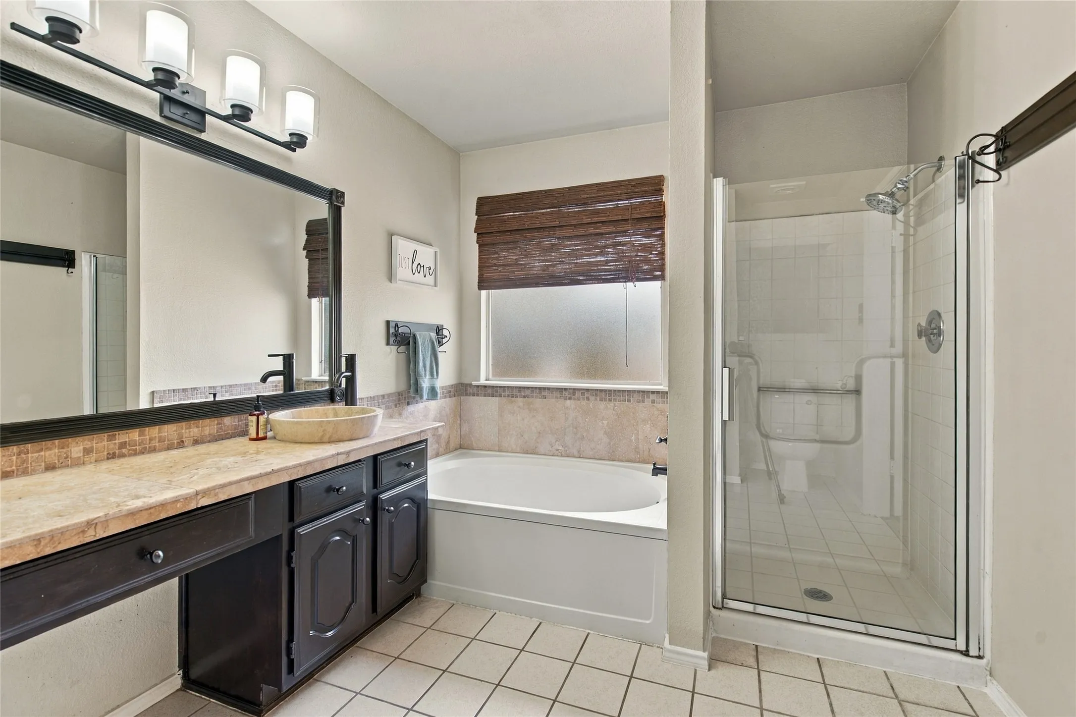 Tiled flooring with a white finish, dark wood vanity with a light-toned countertop, vessel sink with a matte black faucet, built-in bathtub, and a glass-enclosed shower with tile surround