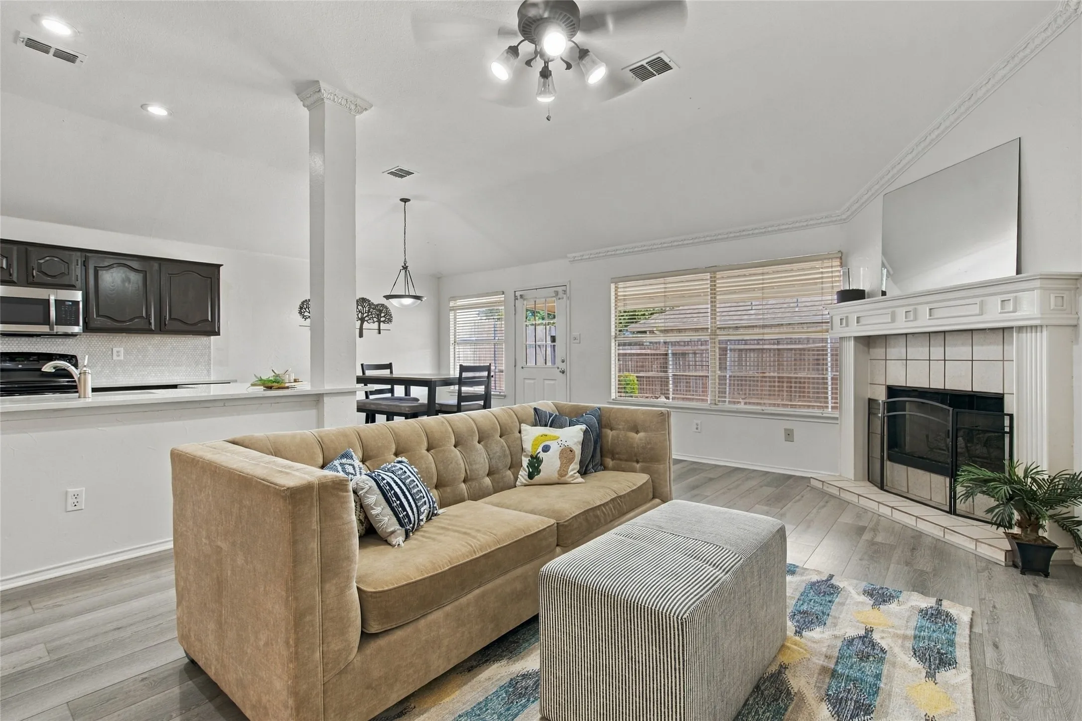 Vaulted ceiling with ceiling fan, wood-finish flooring, white fireplace with tile surround, white crown molding, and kitchen with dark wood cabinetry