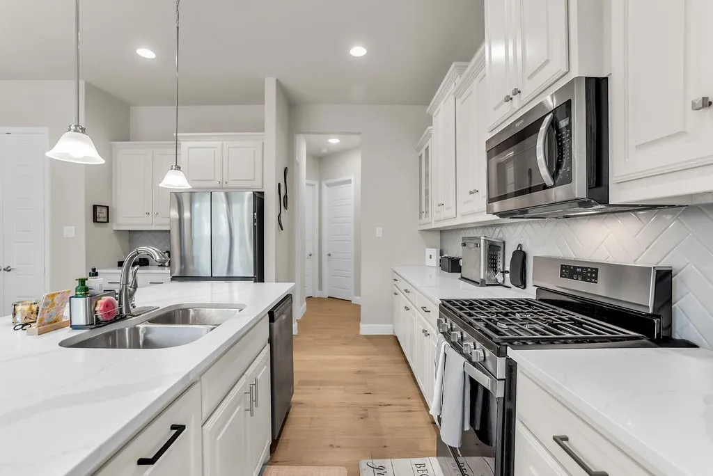 White cabinetry with stainless steel appliances, herringbone tile backsplash, light wood-finish flooring, and a kitchen island with an undermount sink