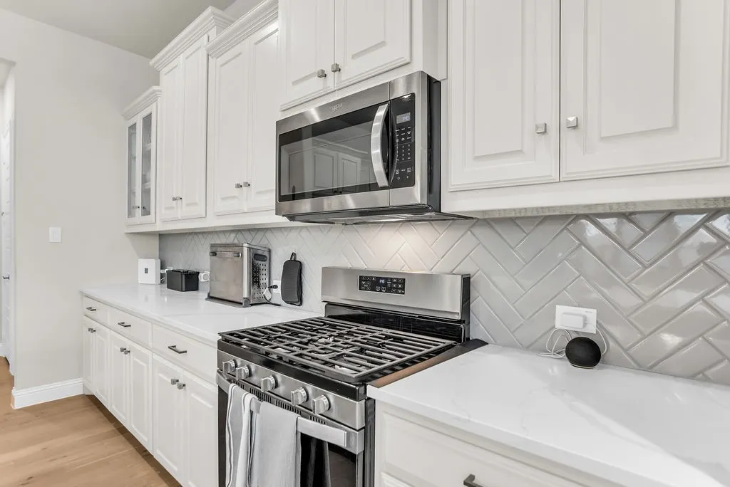 White shaker-style cabinetry, stainless steel gas range, built-in microwave, white herringbone tile backsplash, and light-toned countertops
