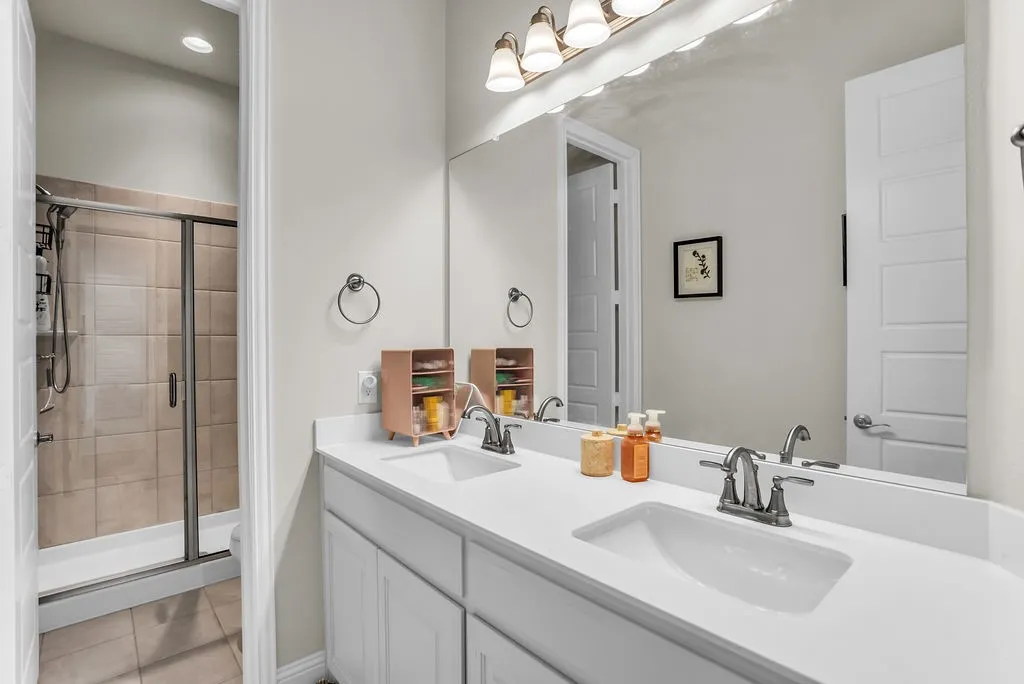Dual vanity with undermount sinks and chrome fixtures, white cabinetry, a large wall-mounted mirror, and a tiled shower with a clear glass enclosure