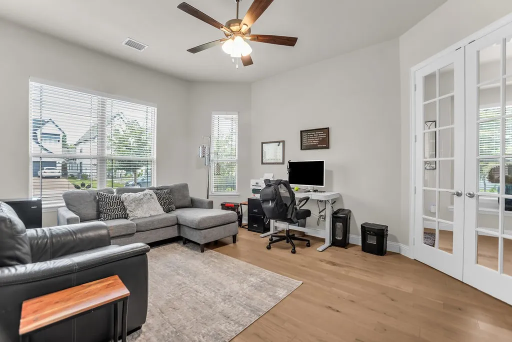 Living area featuring wood-finish flooring, a ceiling fan, multiple windows with blinds, and French doors