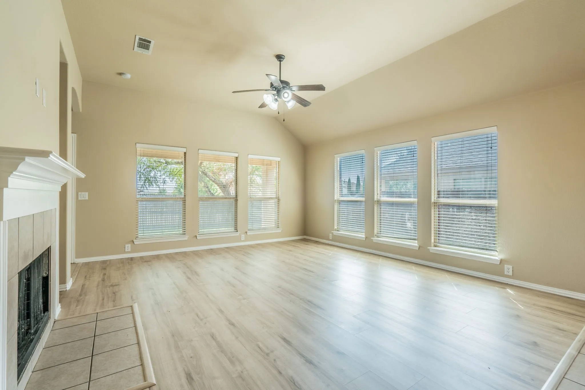 Vaulted ceiling, wood-finish flooring, ceiling fan, multiple windows with blinds, and a tiled fireplace surround