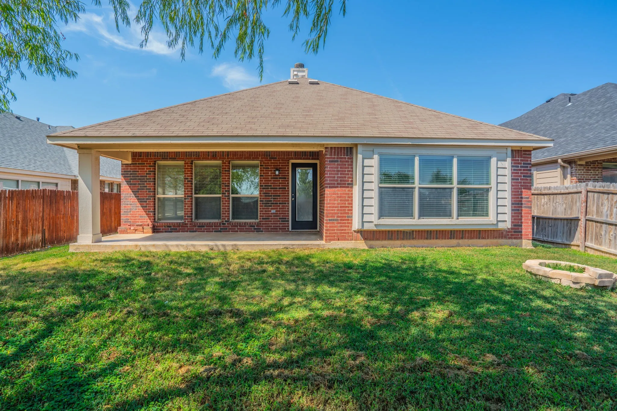 Rear exterior featuring a covered patio, brick facade, shingle roof, and a fenced backyard with a stone fire pit