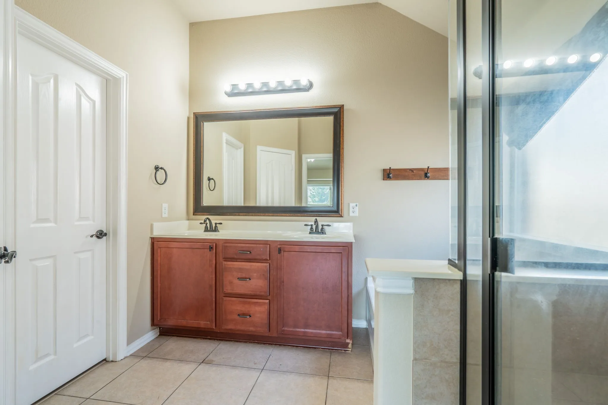 Dual wood-finish vanity with integrated sinks, framed mirror, wall-mounted light fixture, and glass-enclosed shower