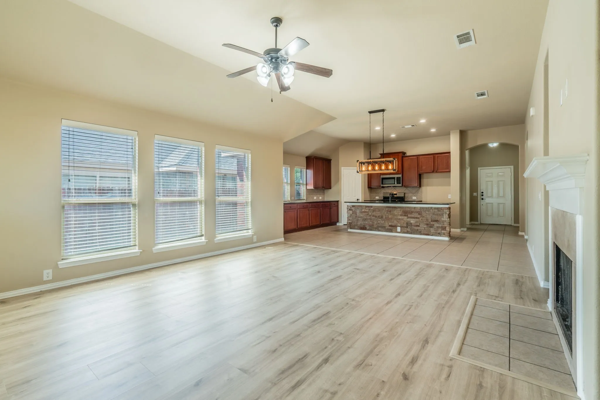 Vaulted ceiling with a ceiling fan, three large windows, wood-finish flooring, a kitchen island with stone detailing, and a fireplace with a mantel