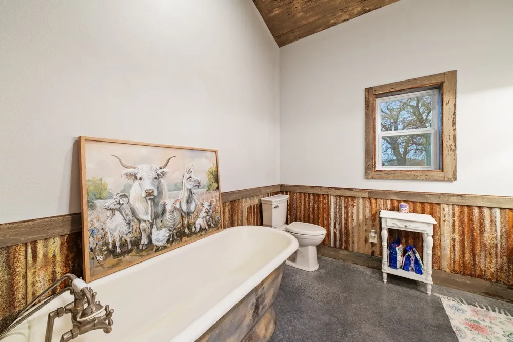 Freestanding bathtub with an antique-style faucet, white toilet, window with wood trim, and corrugated metal wainscoting