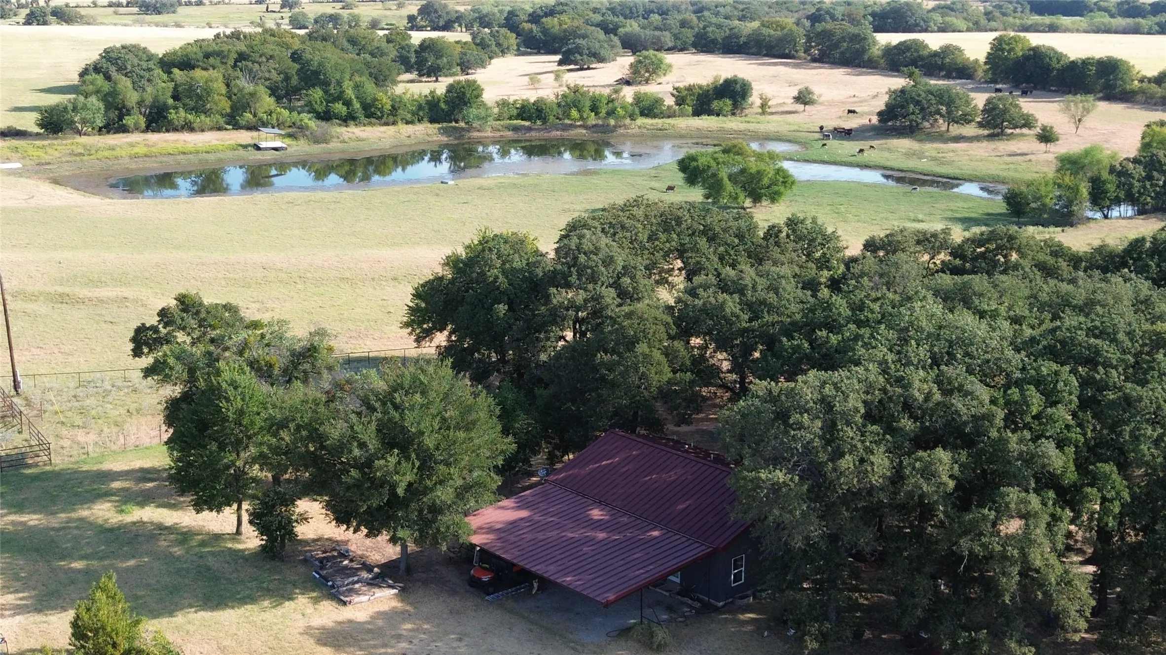 Wooded lot with a red metal roof structure, open fields, and a body of water