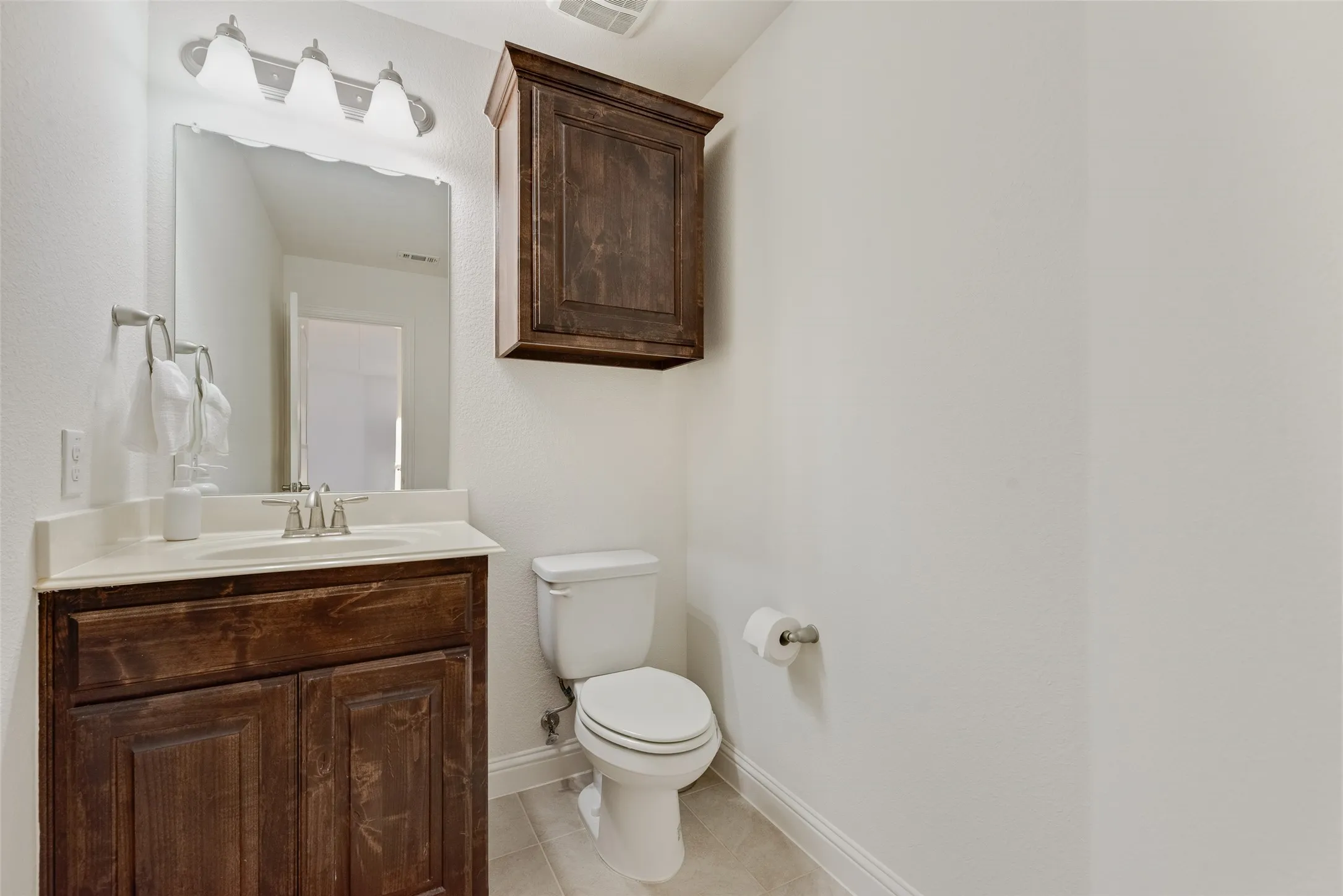 White toilet, dark wood-finish vanity with an integrated sink, wall-mounted mirror, and overhead tri-light fixture