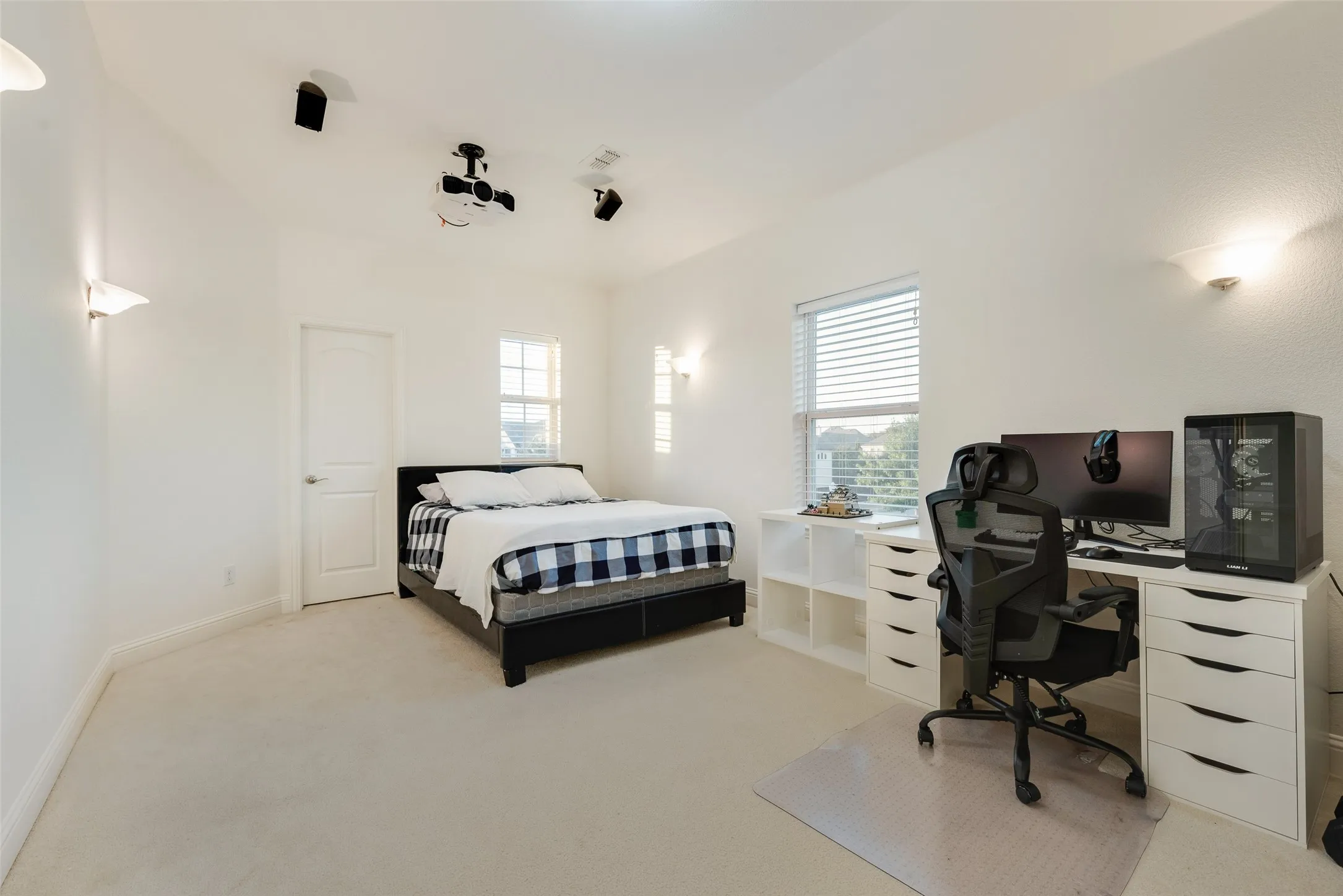Recessed ceiling-mounted projector, integrated ceiling speakers, dual windows with blinds, wall-mounted sconce lighting, and soft carpet flooring