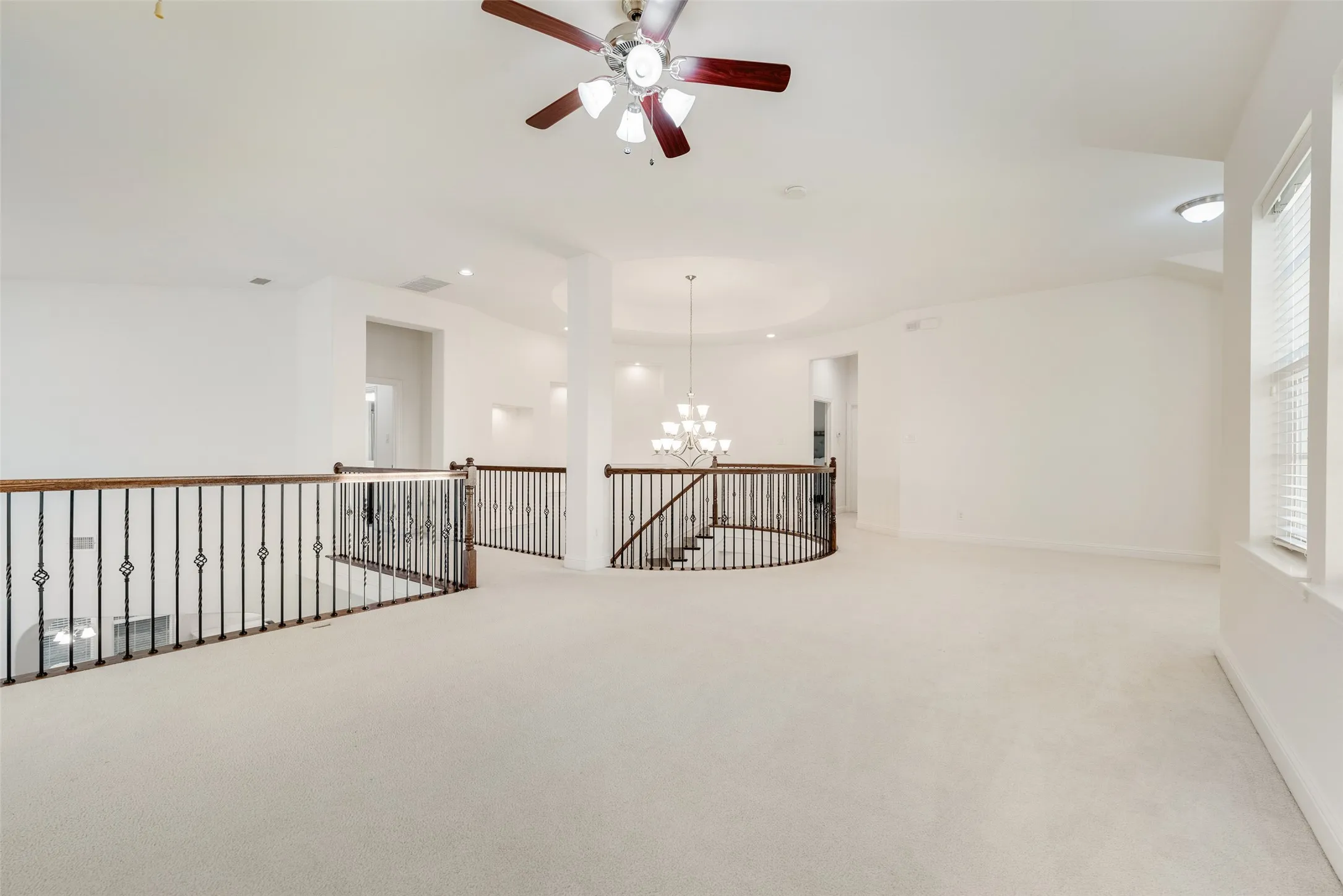 Expansive carpeted loft area featuring an ornate chandelier, recessed lighting, a ceiling fan, and decorative wrought iron and wood railings