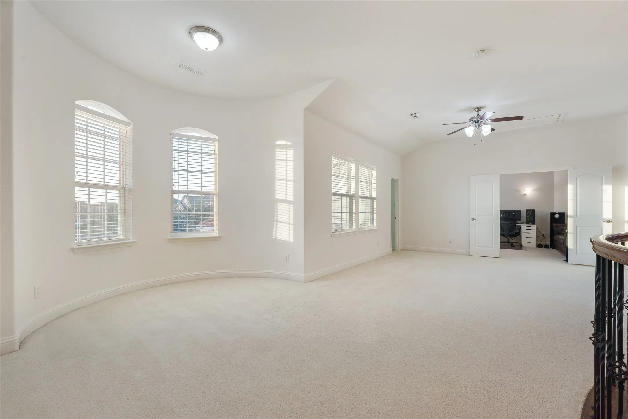 Spacious carpeted room featuring a vaulted ceiling with a ceiling fan, multiple windows with blinds, and a built-in archway leading to an additional space