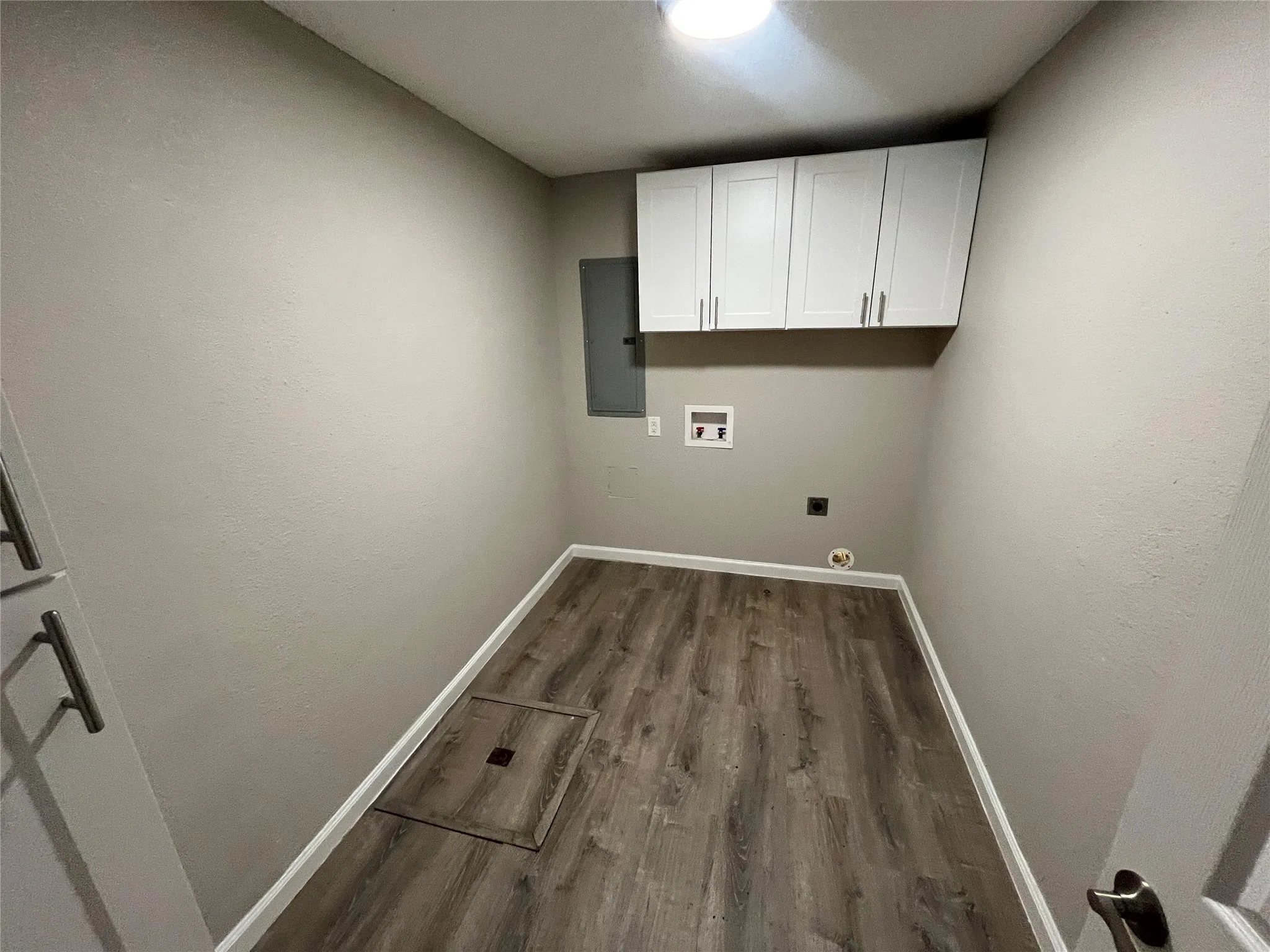 Laundry area featuring wood-finish flooring, overhead cabinetry with silver-tone pulls, wall-mounted electrical panel, and recessed utility connections