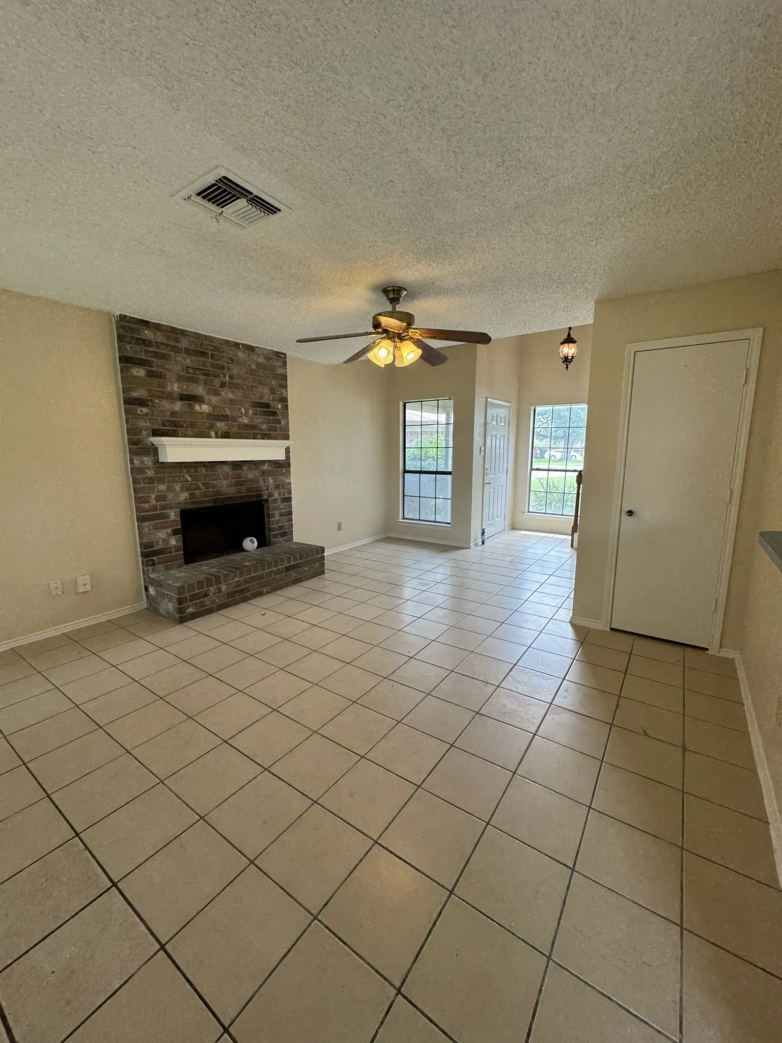 Spacious room featuring neutral tile flooring, a brick fireplace with a white mantel, multiple windows, and a ceiling fan