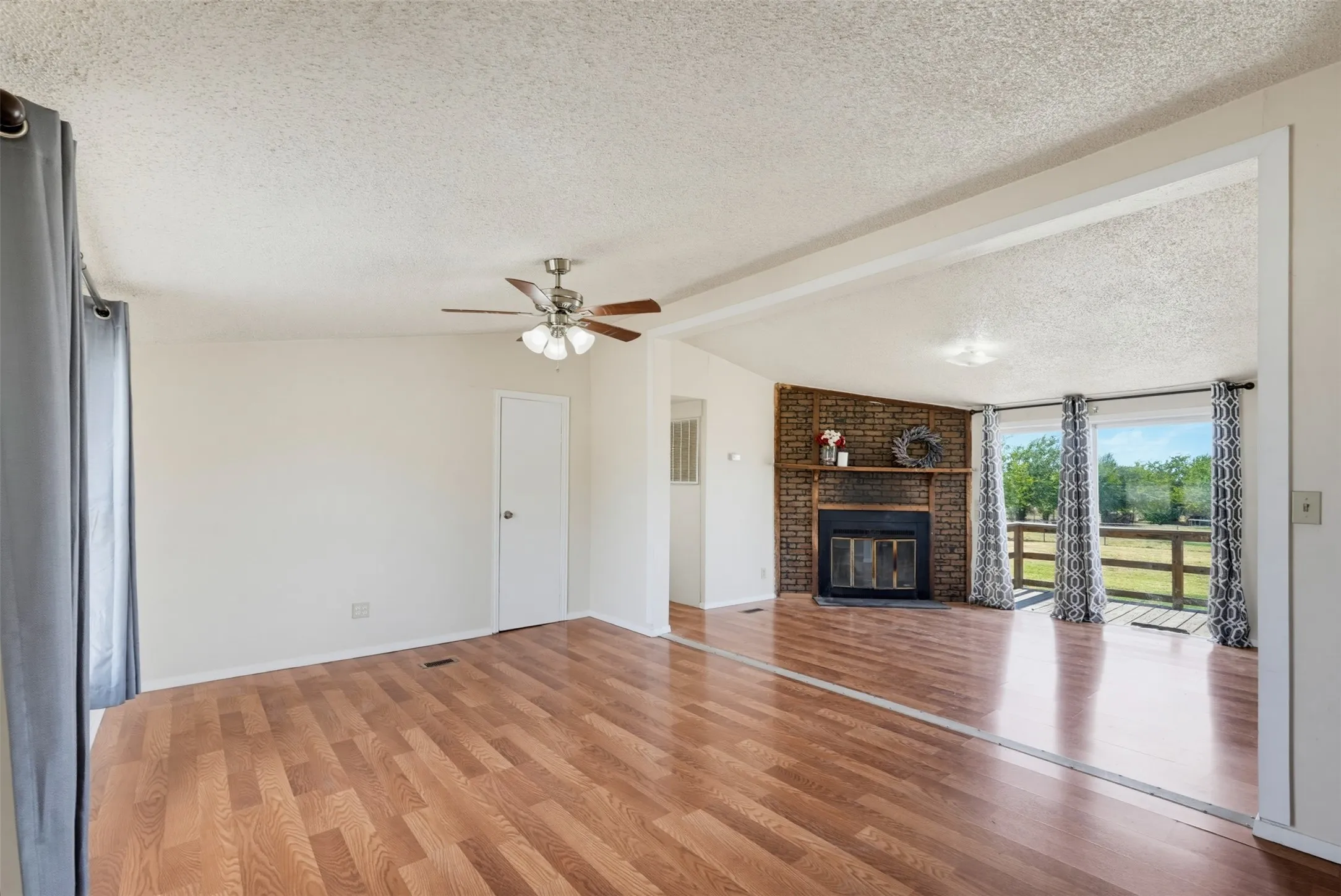 Wood-finish flooring throughout, ceiling fan with light fixture, brick fireplace with mantel, and sliding glass doors