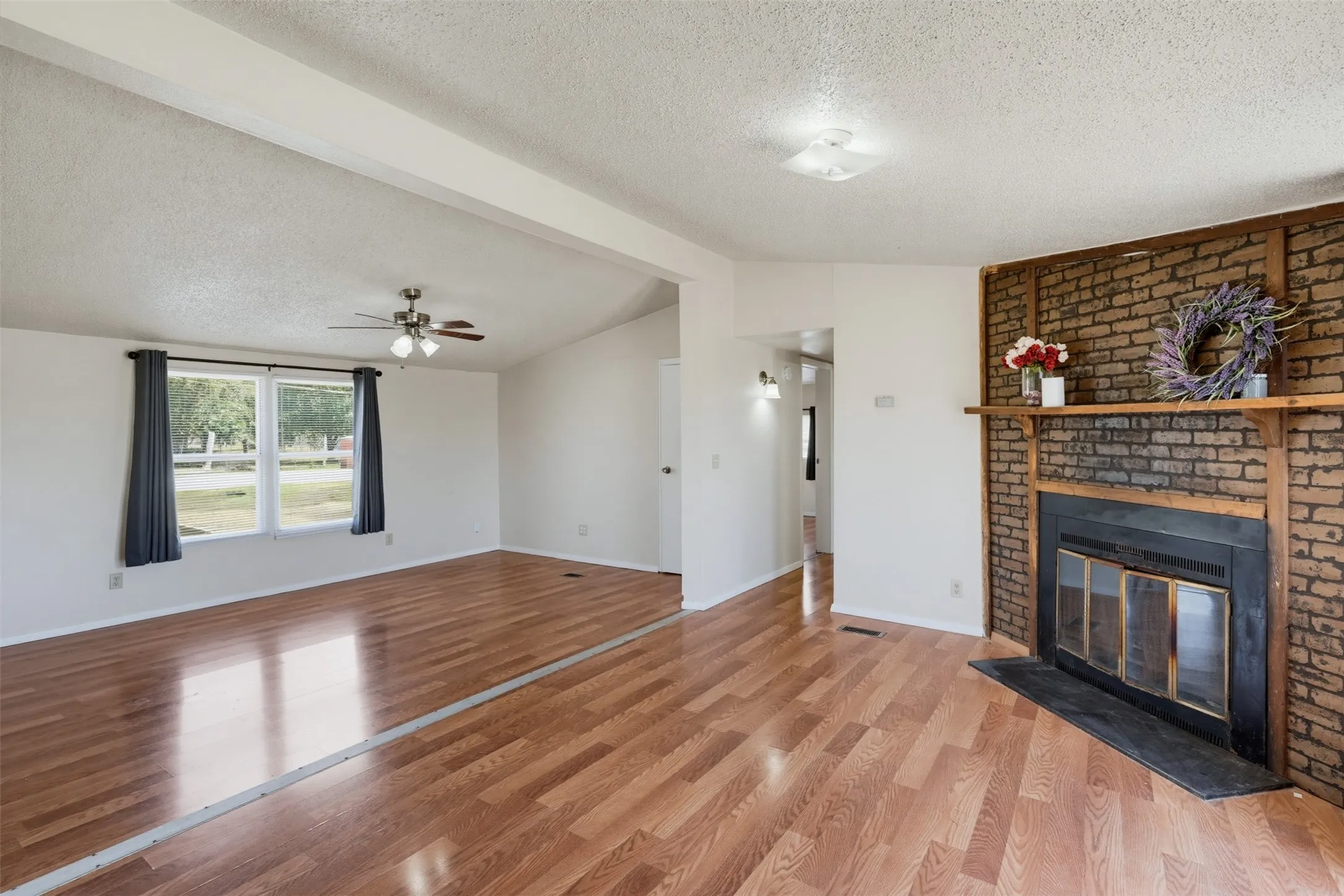 Vaulted ceiling with exposed beam, wood-finish flooring, brick fireplace with wood mantel, and large windows with outdoor scenery