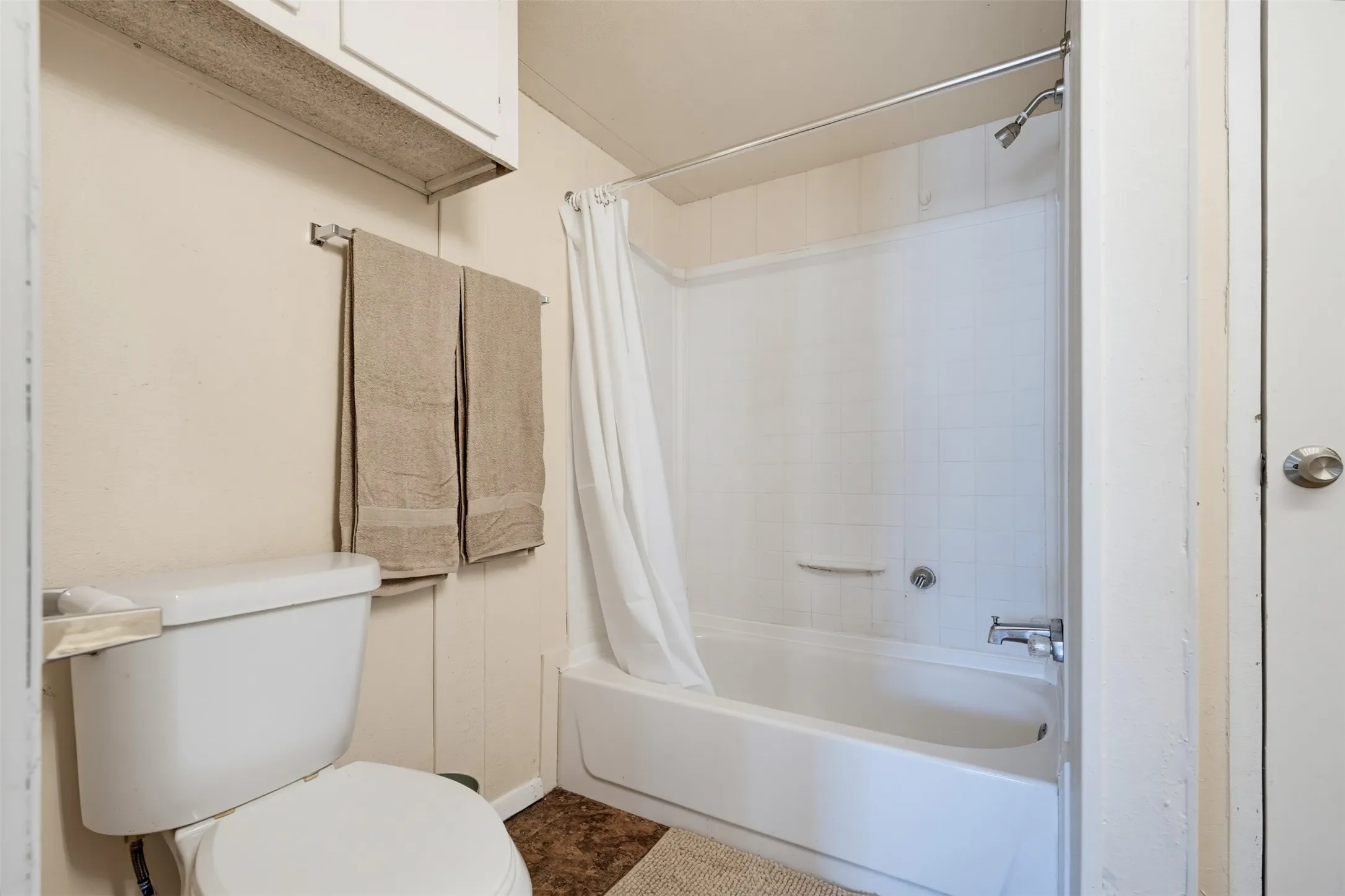 White toilet with an overhead storage cabinet, chrome towel bar, and a tub-shower combination with a white shower curtain