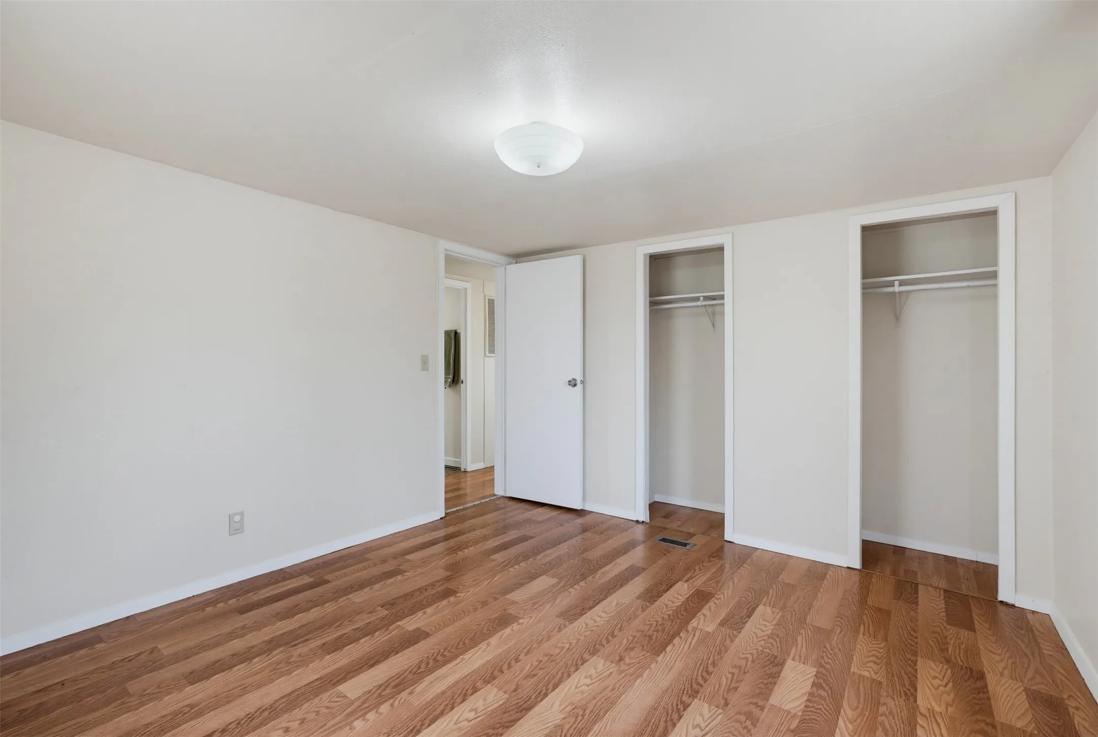Wood-finish flooring throughout, bright white walls, two open closet spaces with hanging rods, and an overhead flush-mount light fixture