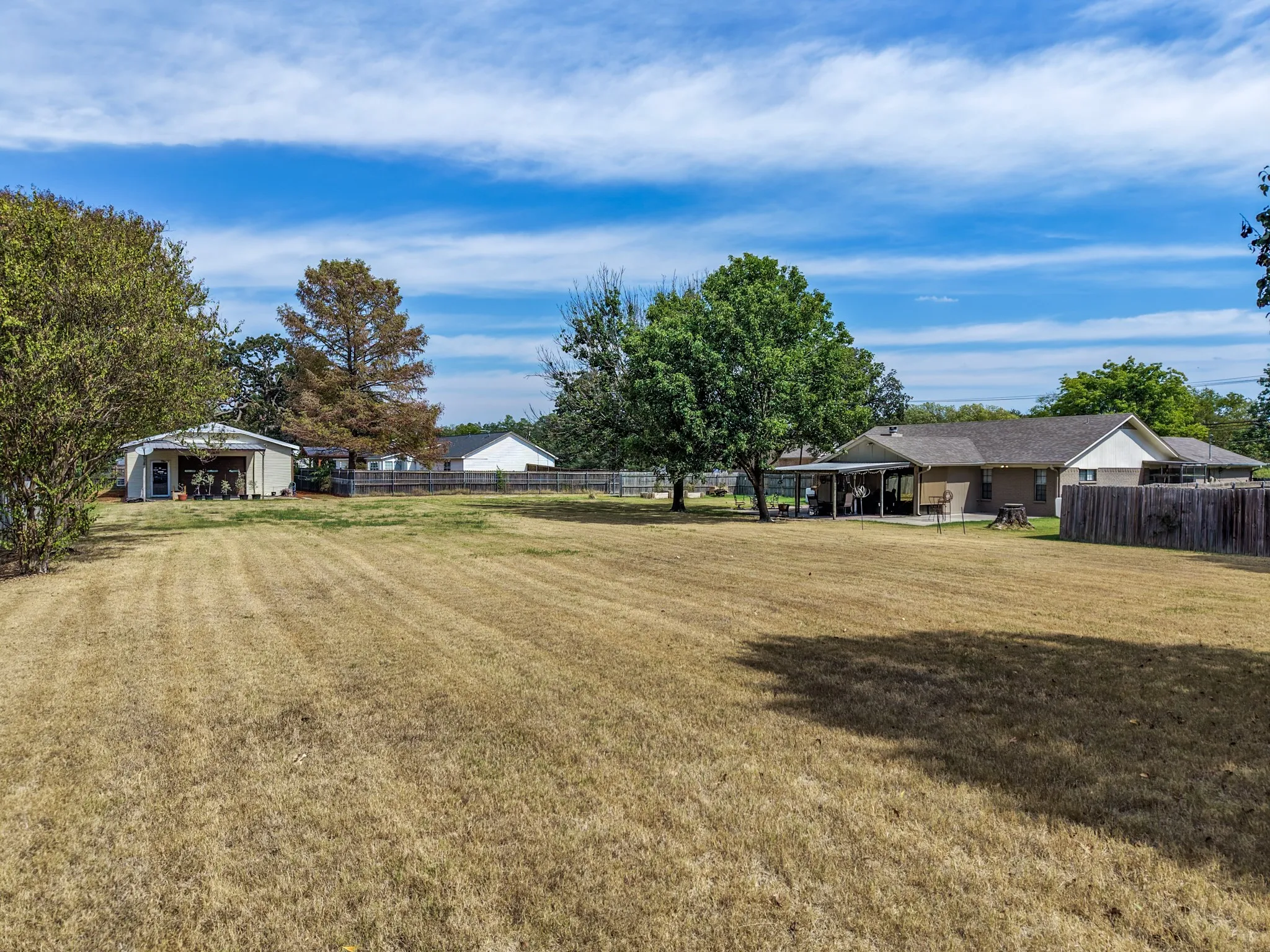 Expansive grassy area with mature trees, a detached garage, and a single-story residence featuring an extended patio with an overhead cover