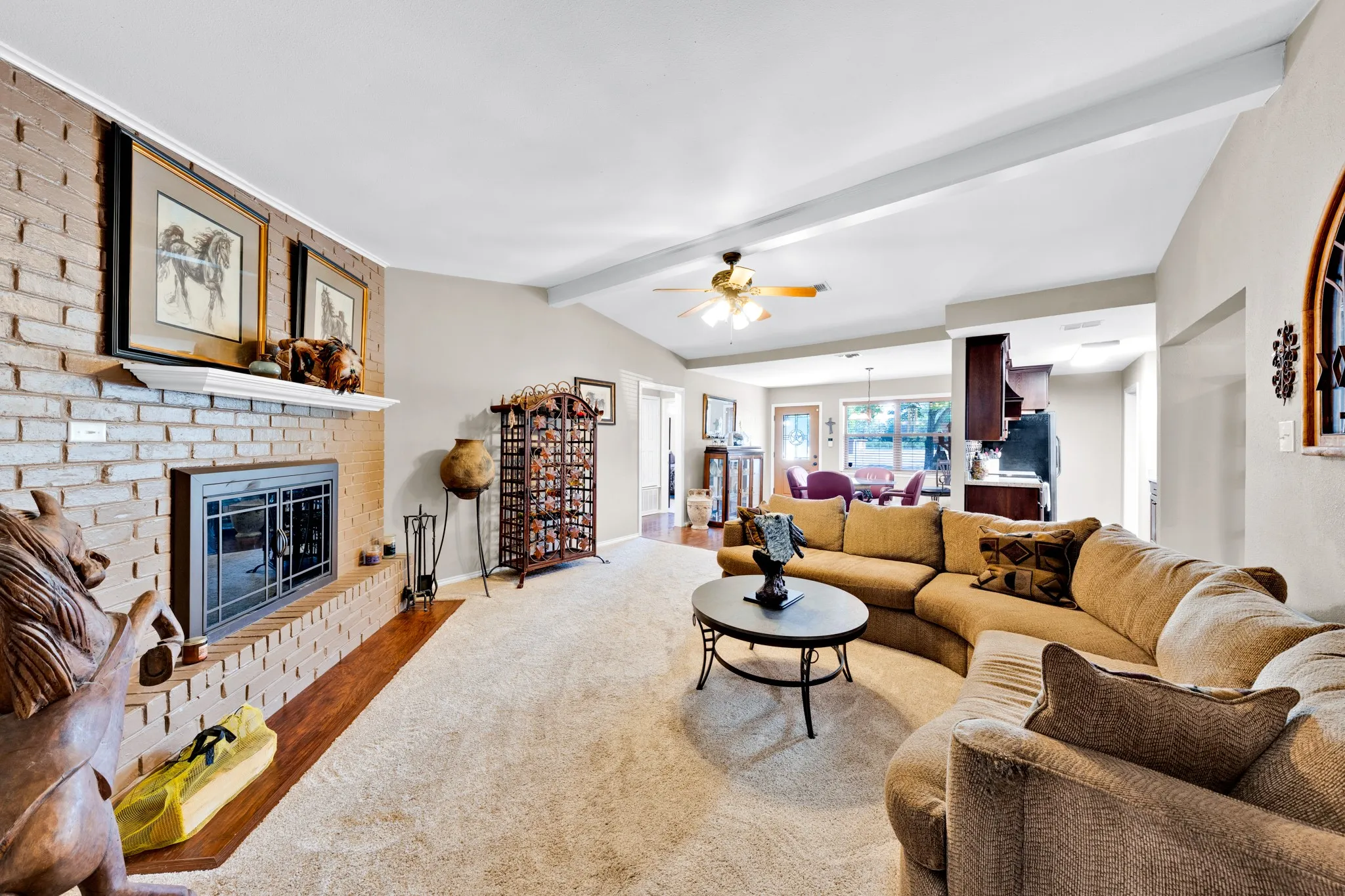 Brick fireplace with a wood mantel, vaulted ceiling with exposed beams, ceiling fan, and carpet flooring