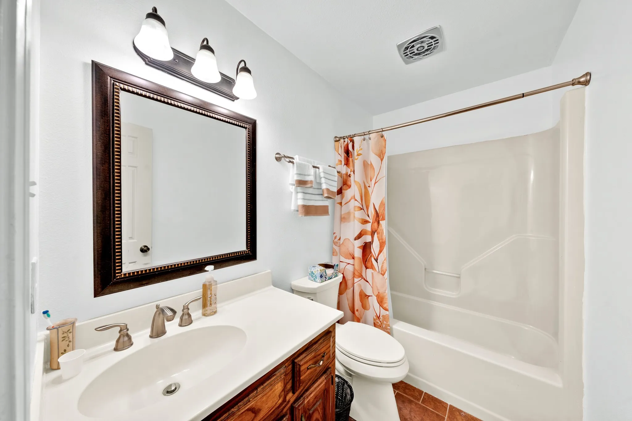 White vanity with an integrated sink and bronze-finish faucet, framed mirror with a three-light fixture, and a bath/shower combination with a bronze-finish shower rod