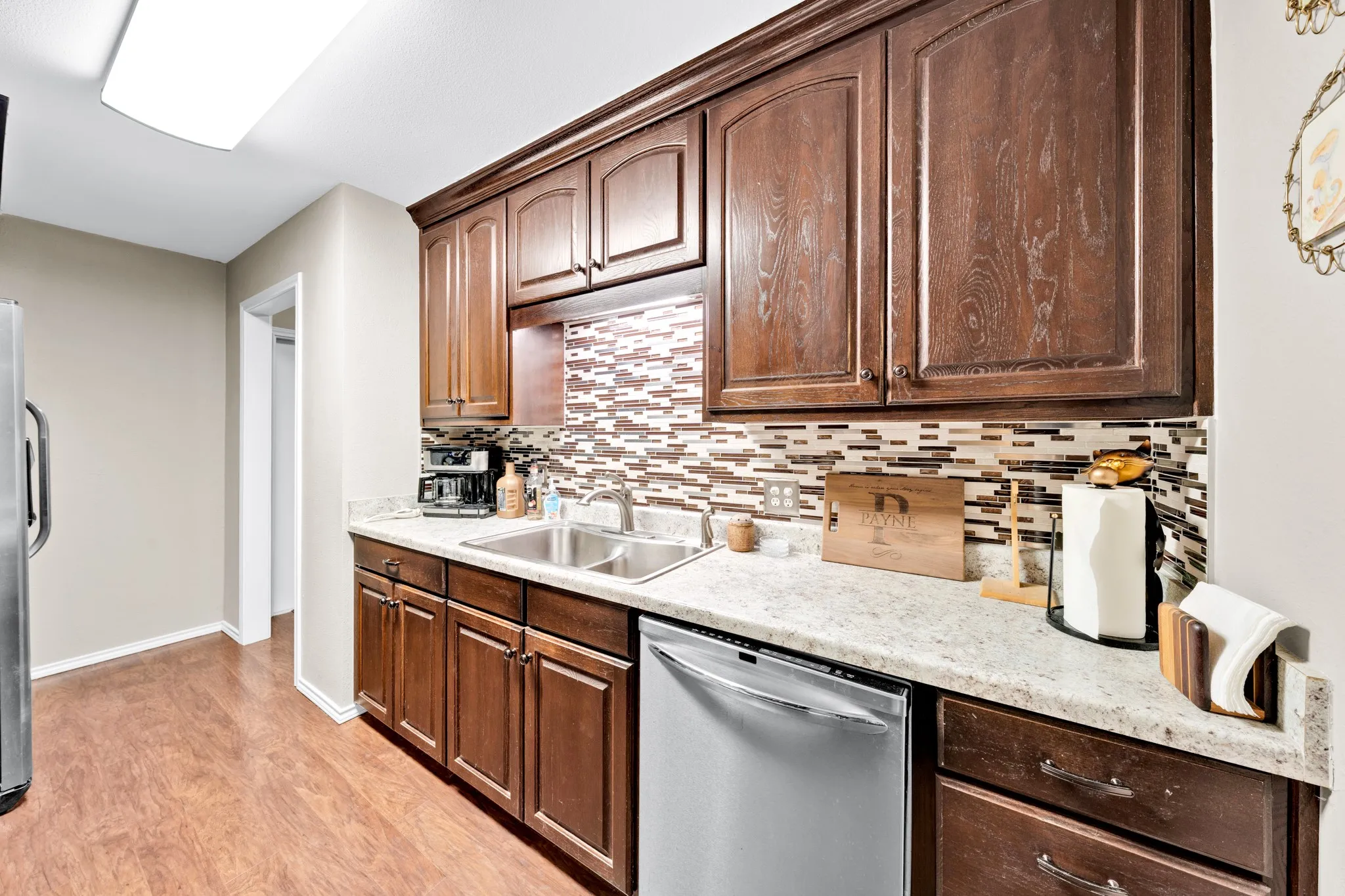 Wood-finish flooring, dark wood cabinetry, light-toned countertops, mosaic tile backsplash, and a double basin sink