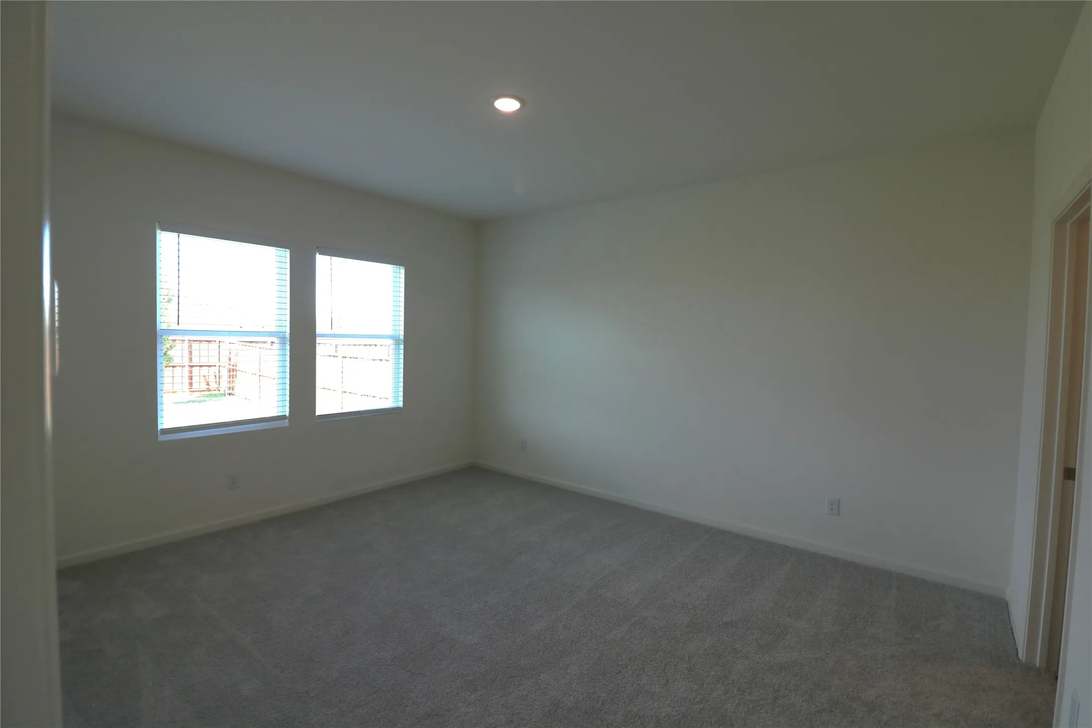 Recessed ceiling lighting illuminates a spacious room featuring two windows with blinds and neutral carpeting