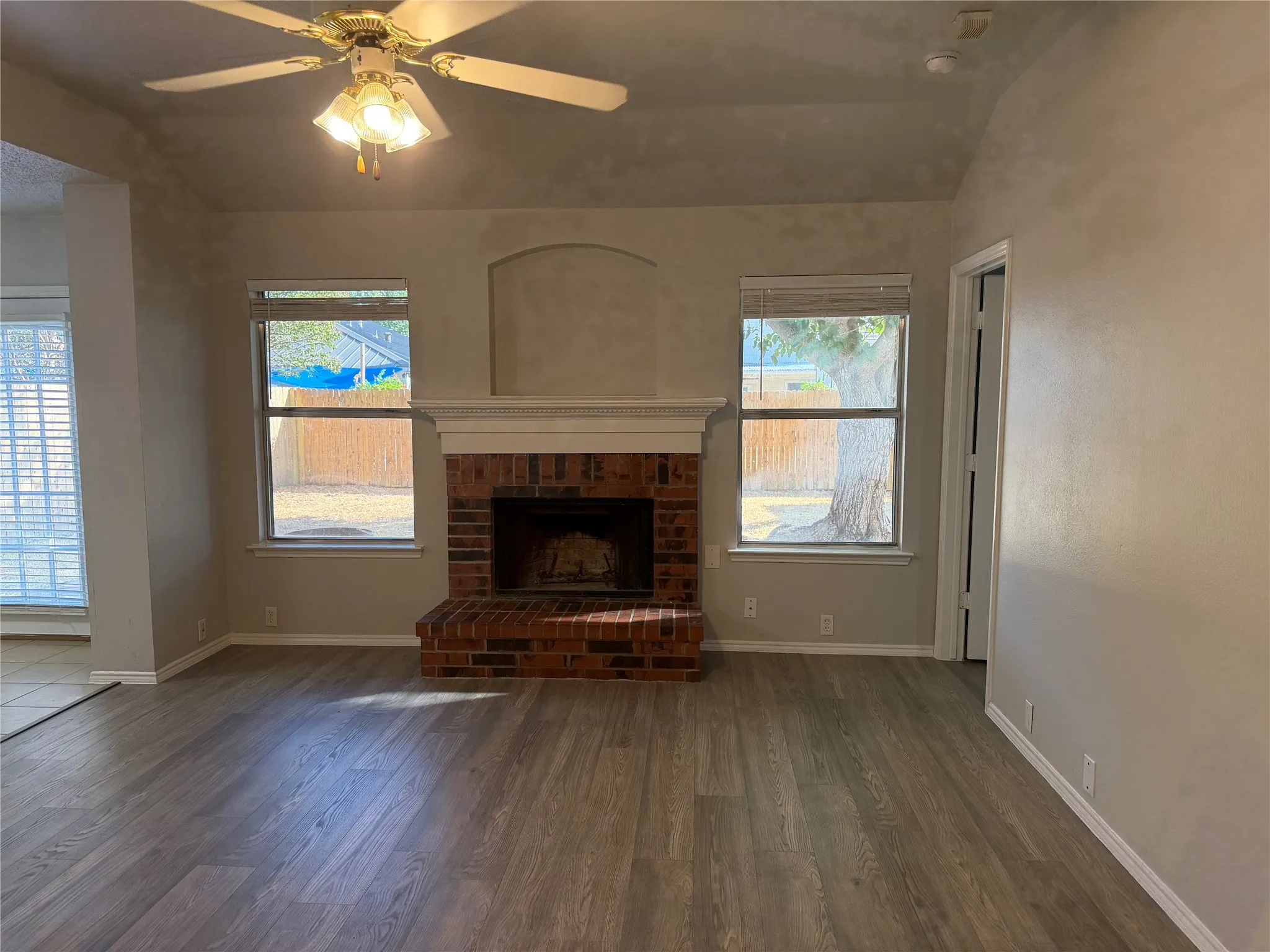 Vaulted ceiling with a ceiling fan, brick fireplace with a mantel, wood-finish flooring, and multiple windows