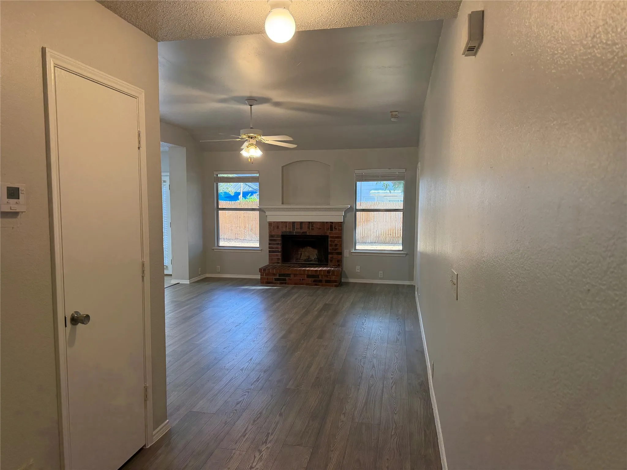 Wood-finish flooring throughout, brick fireplace with mantel, two windows, and a ceiling fan