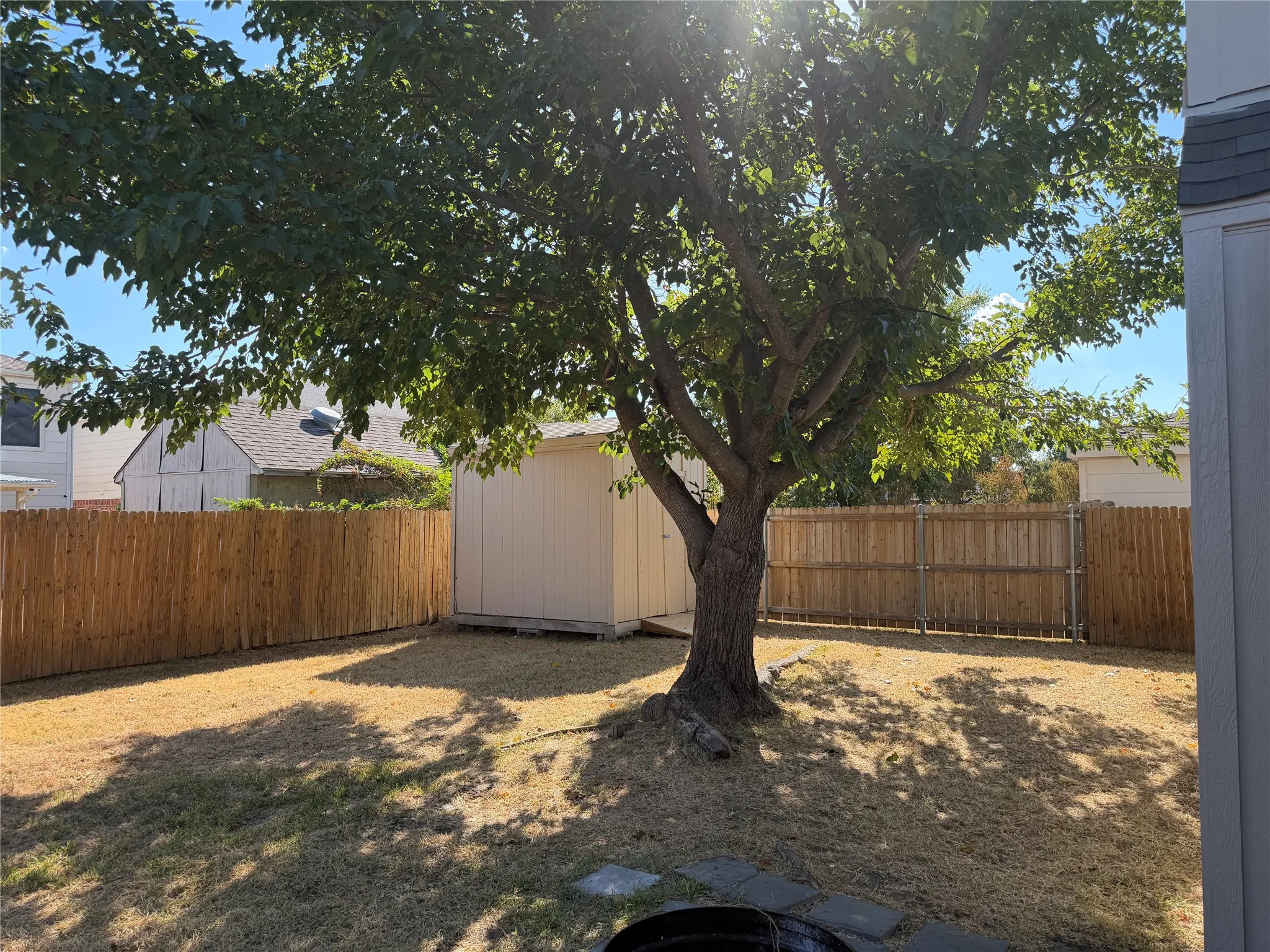 Fenced backyard featuring a mature shade tree, a storage shed, and stone paving