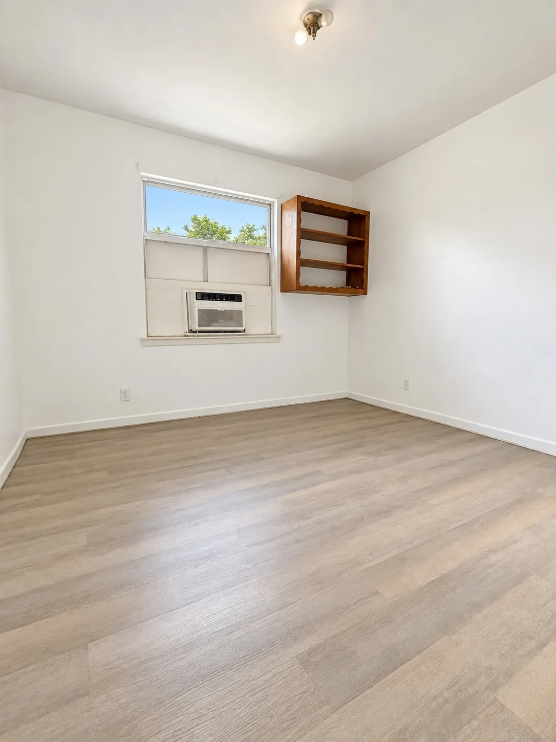 Light wood-finish flooring, a window-mounted air conditioning unit, built-in shelving, and a flush-mount ceiling light