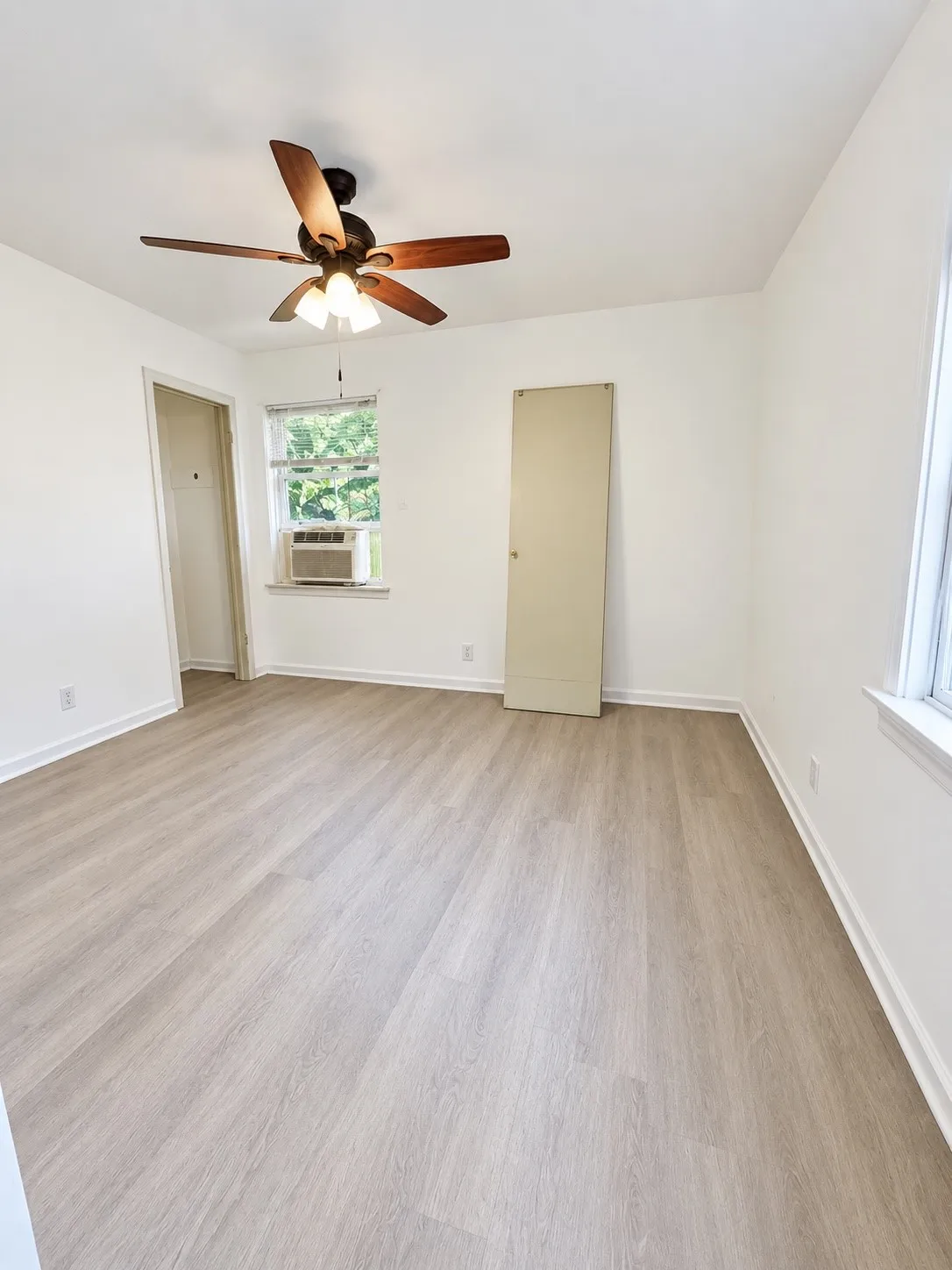 Wood-finish flooring, ceiling fan with integrated lighting, white walls, and a window unit air conditioner