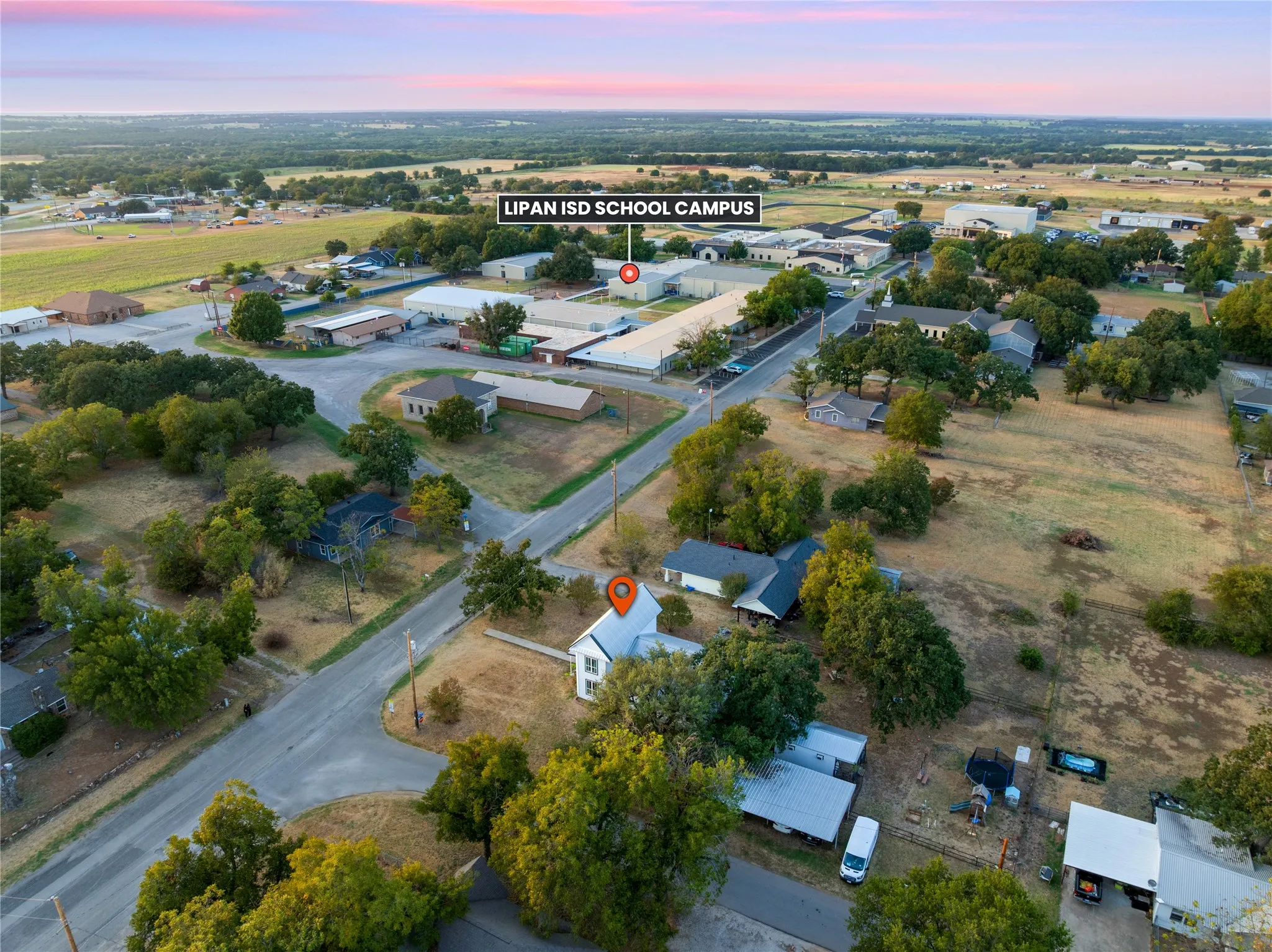 Aerial view of the property and surrounding neighborhood, featuring a two-story white structure with a dark roof and mature trees on a lot with a natural grass surface