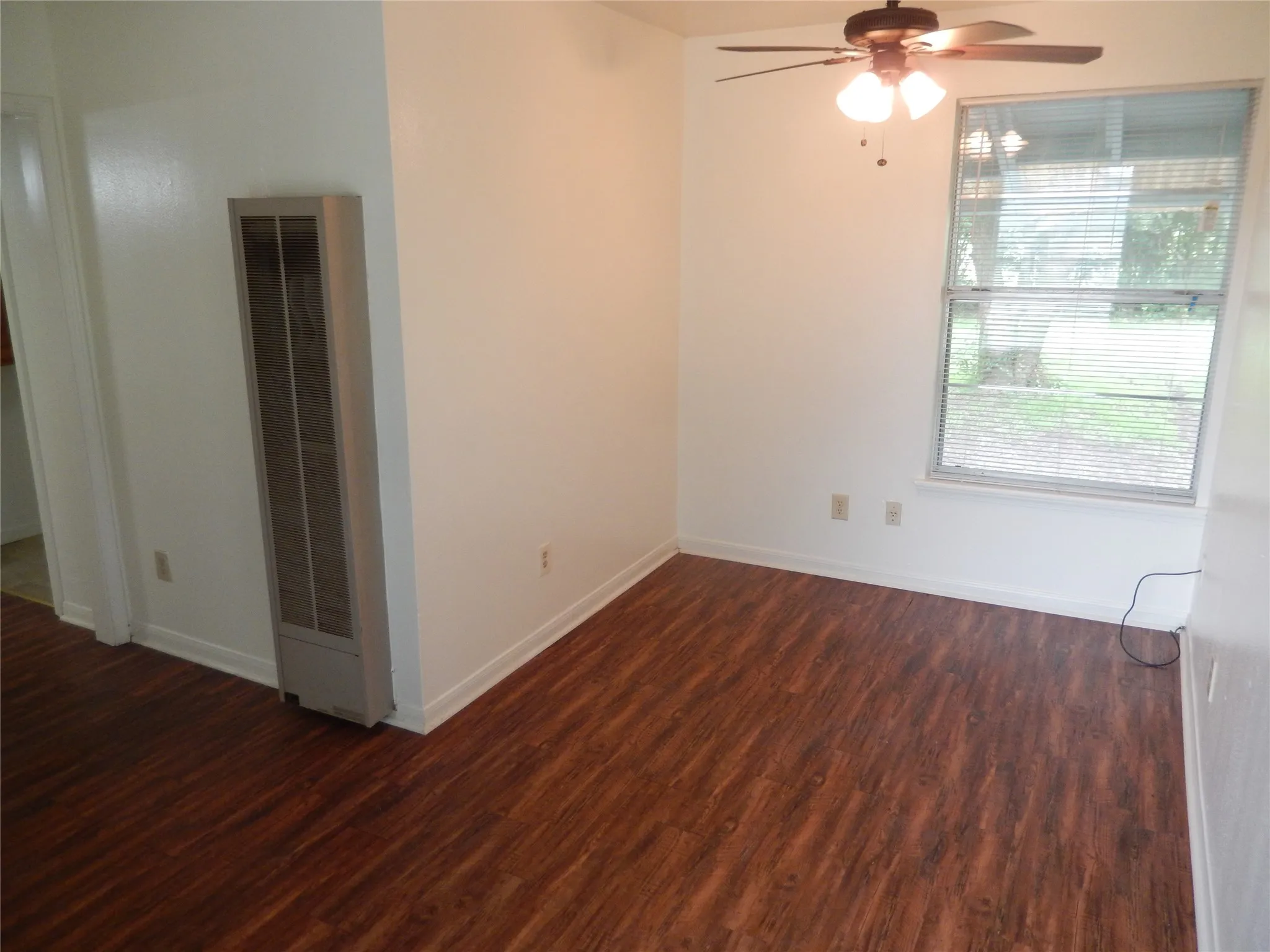 Wood-finish flooring throughout, white walls, ceiling fan with light fixture, window with horizontal blinds, and a permanent wall-mounted heater