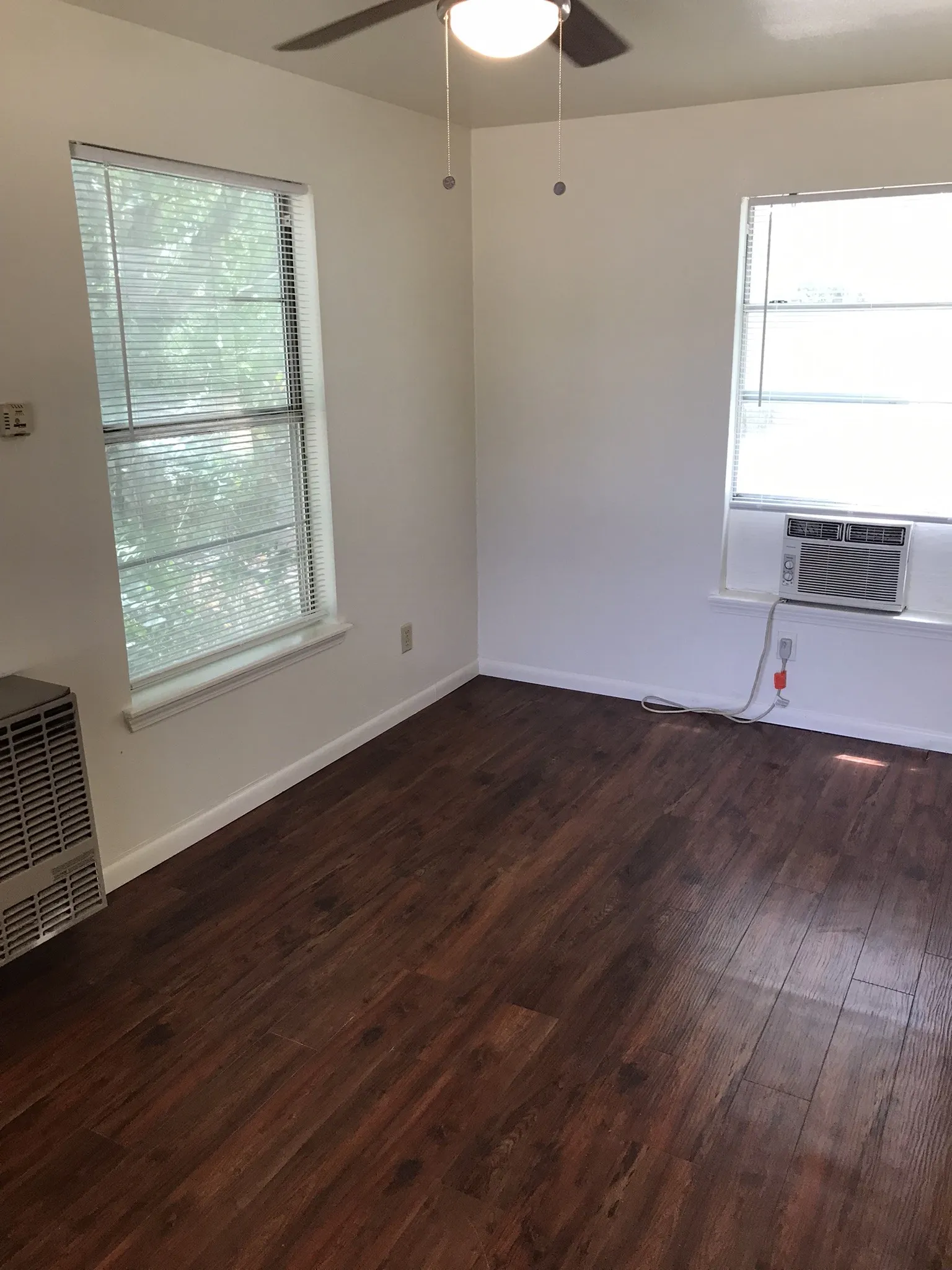Wood-finish flooring throughout, two windows with blinds, ceiling fan, wall-mounted heating unit, and window-mounted air conditioning unit