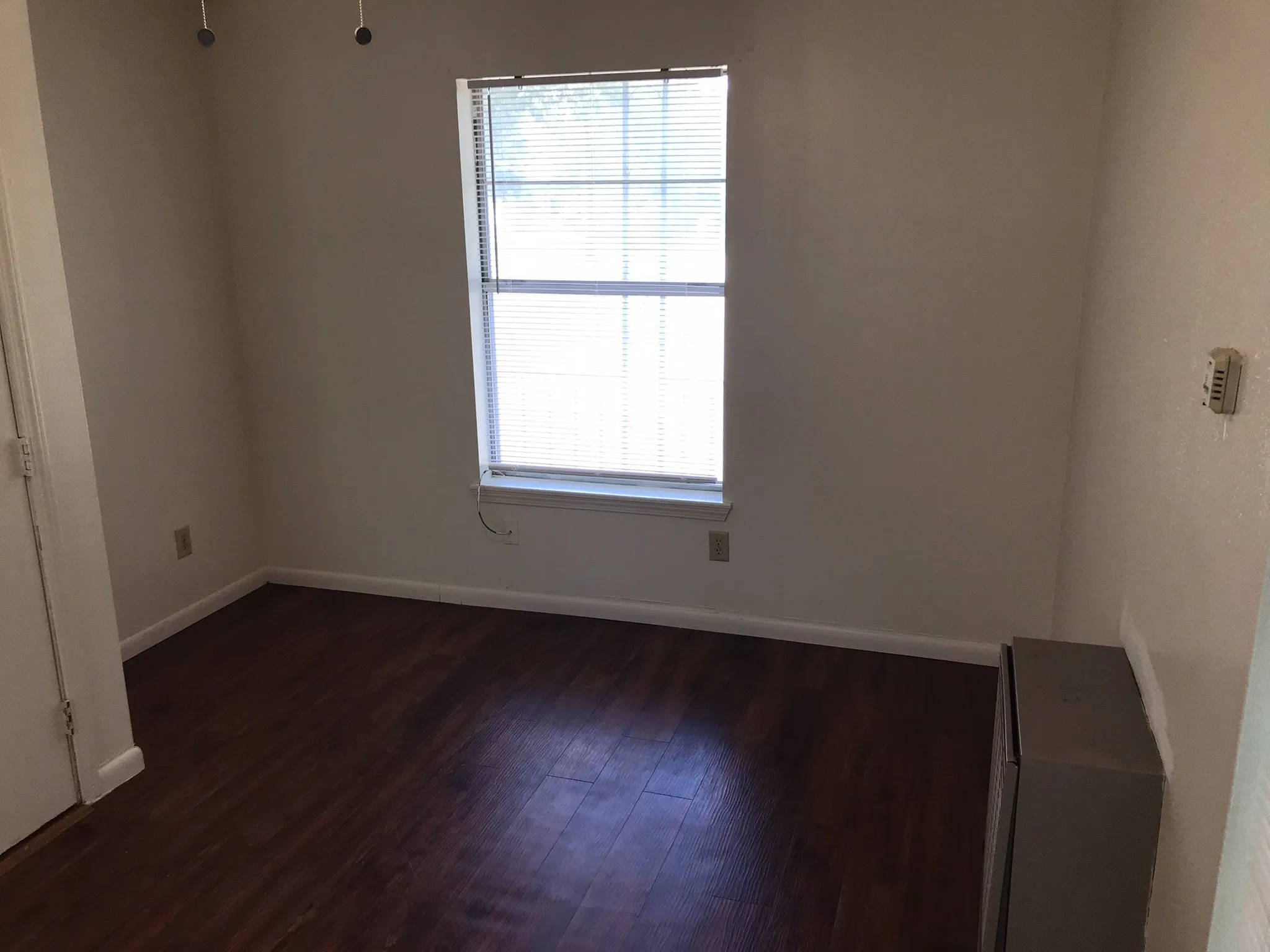 Wood-finish flooring with white baseboards, a single window, and a wall-mounted thermostat