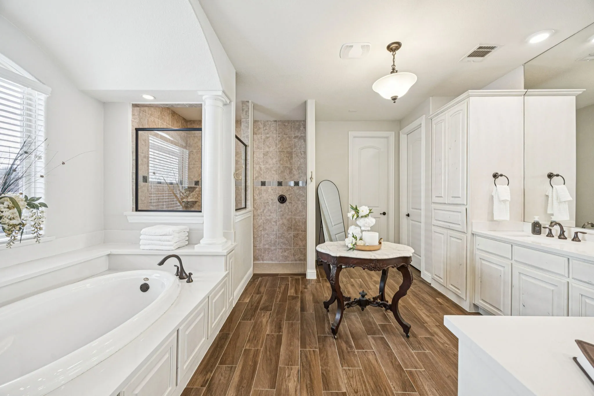 Soaking tub with dark bronze-finish fixtures, a separate glass-enclosed shower with tiled walls, dual vanity with white cabinetry and countertop, wood-finish flooring, and a decorative column