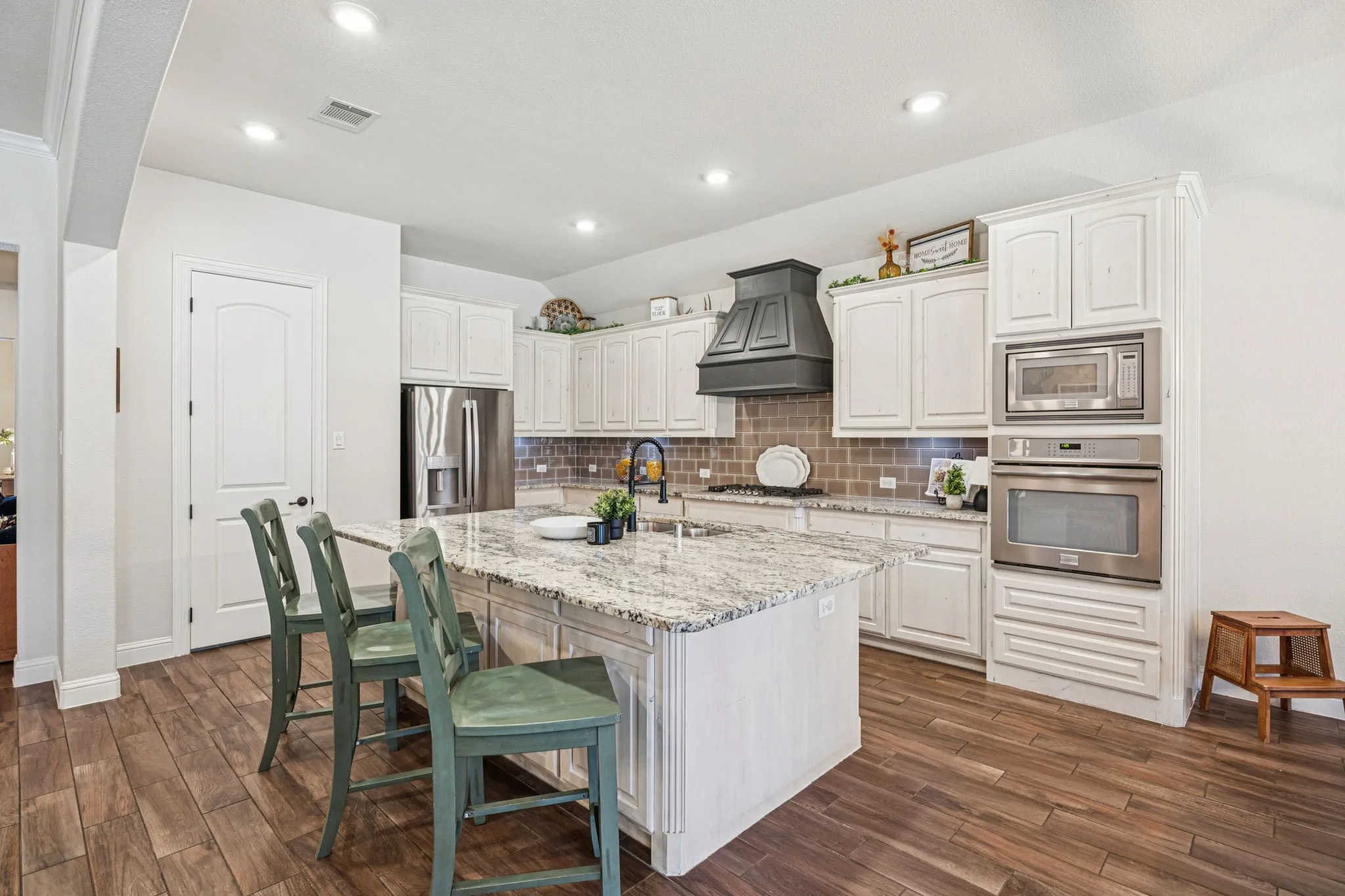 Central island with a granite countertop, white cabinetry with a built-in oven and microwave, wood-finish flooring, and a subway tile backsplash