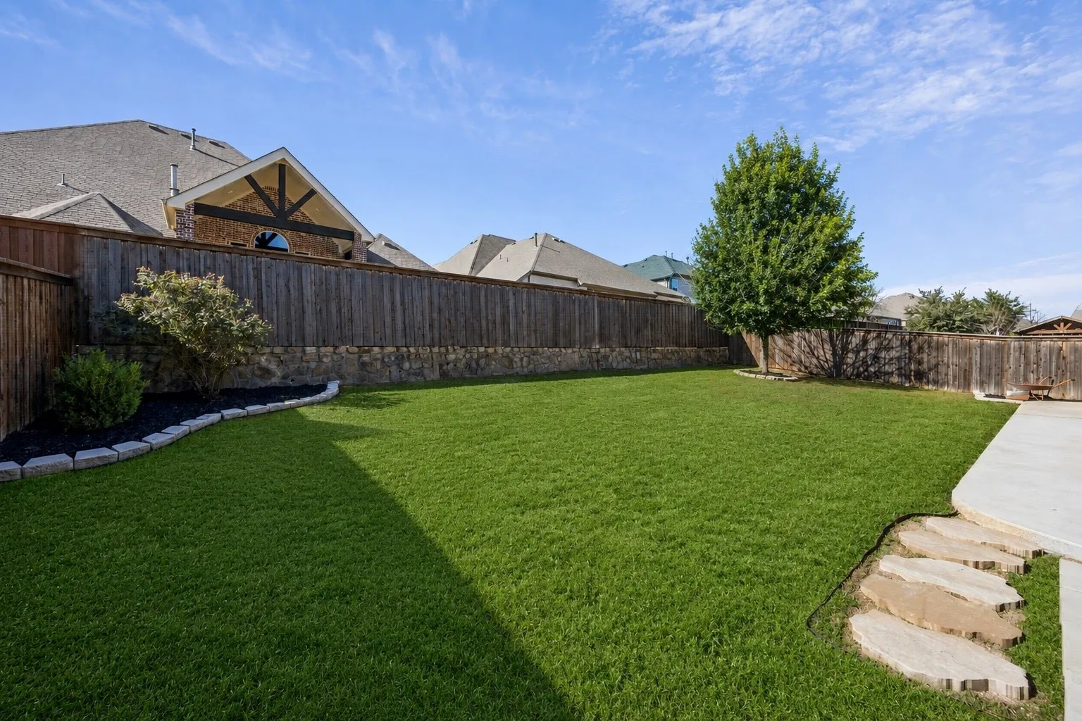 Expansive green lawn, wood privacy fence with stone base, mature trees, concrete patio, and natural stone stepping pavers