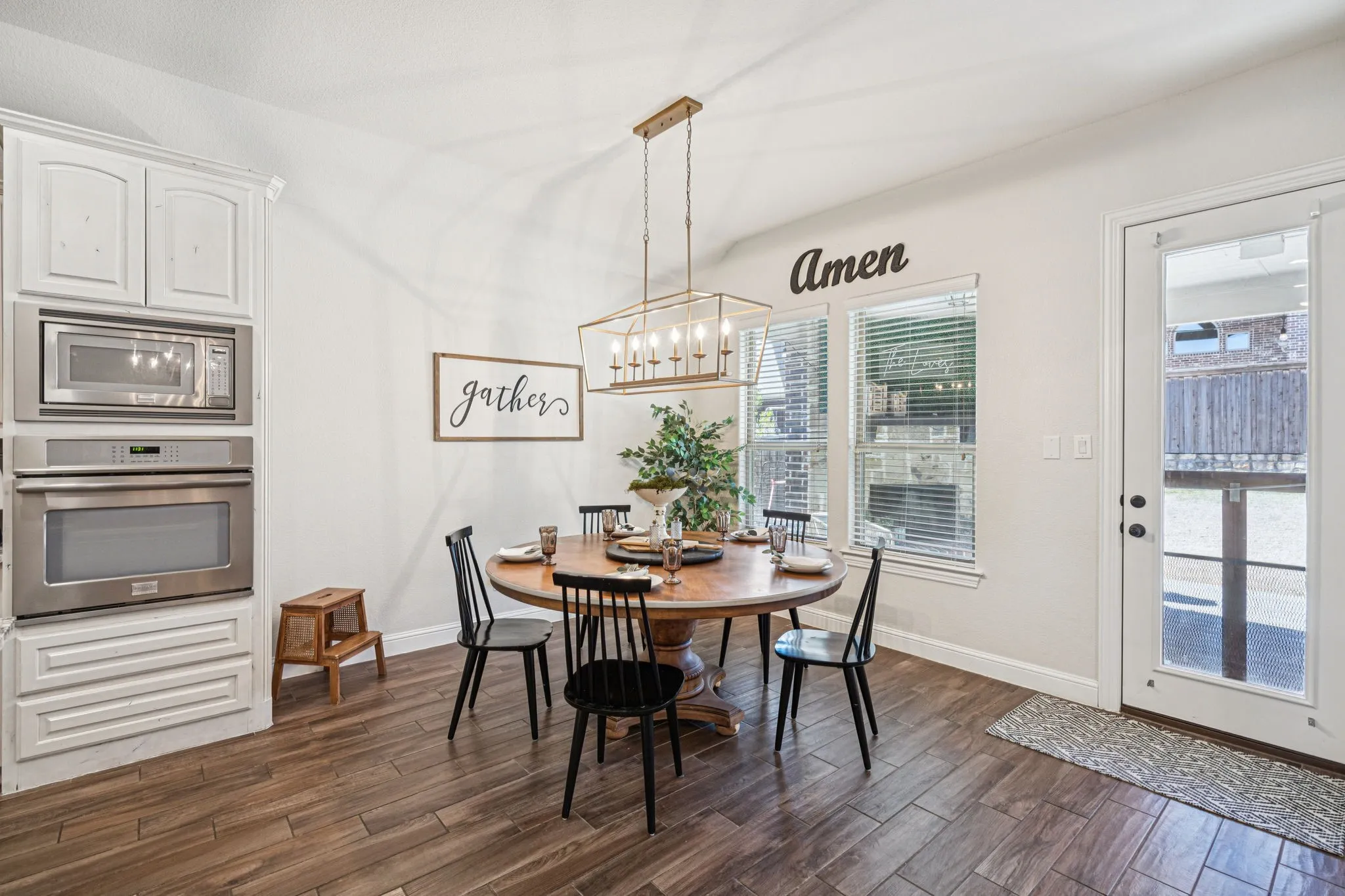 Built-in stainless steel double oven, white cabinetry with crown molding, wood-finish flooring, two large windows with blinds, and a glass-paneled exterior door