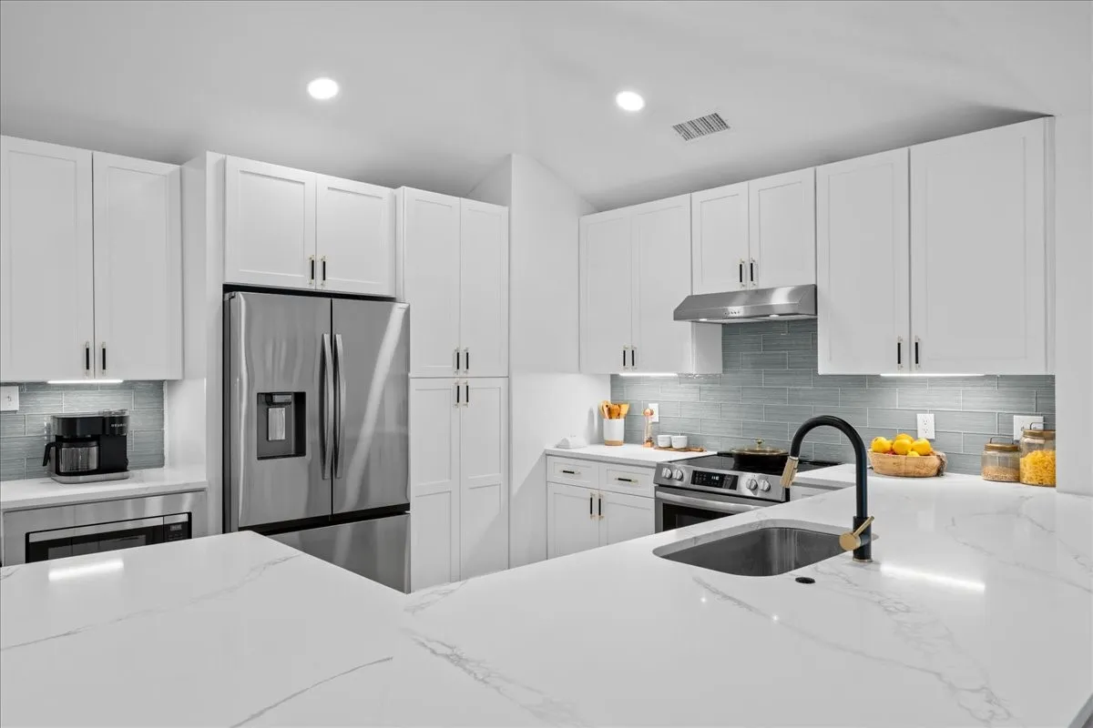 White shaker cabinetry with modern hardware, stainless steel appliances, a large single-basin sink with a matte black faucet, light-toned countertops, and a full-height tile backsplash