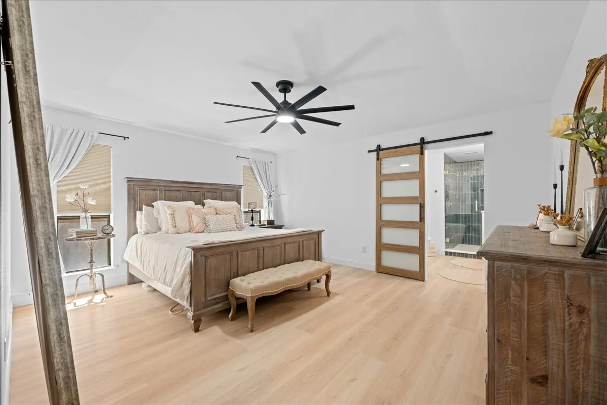 Wood-finish flooring, ceiling fan with integrated lighting, two windows with window treatments, and a wood-panel and frosted-glass barn door