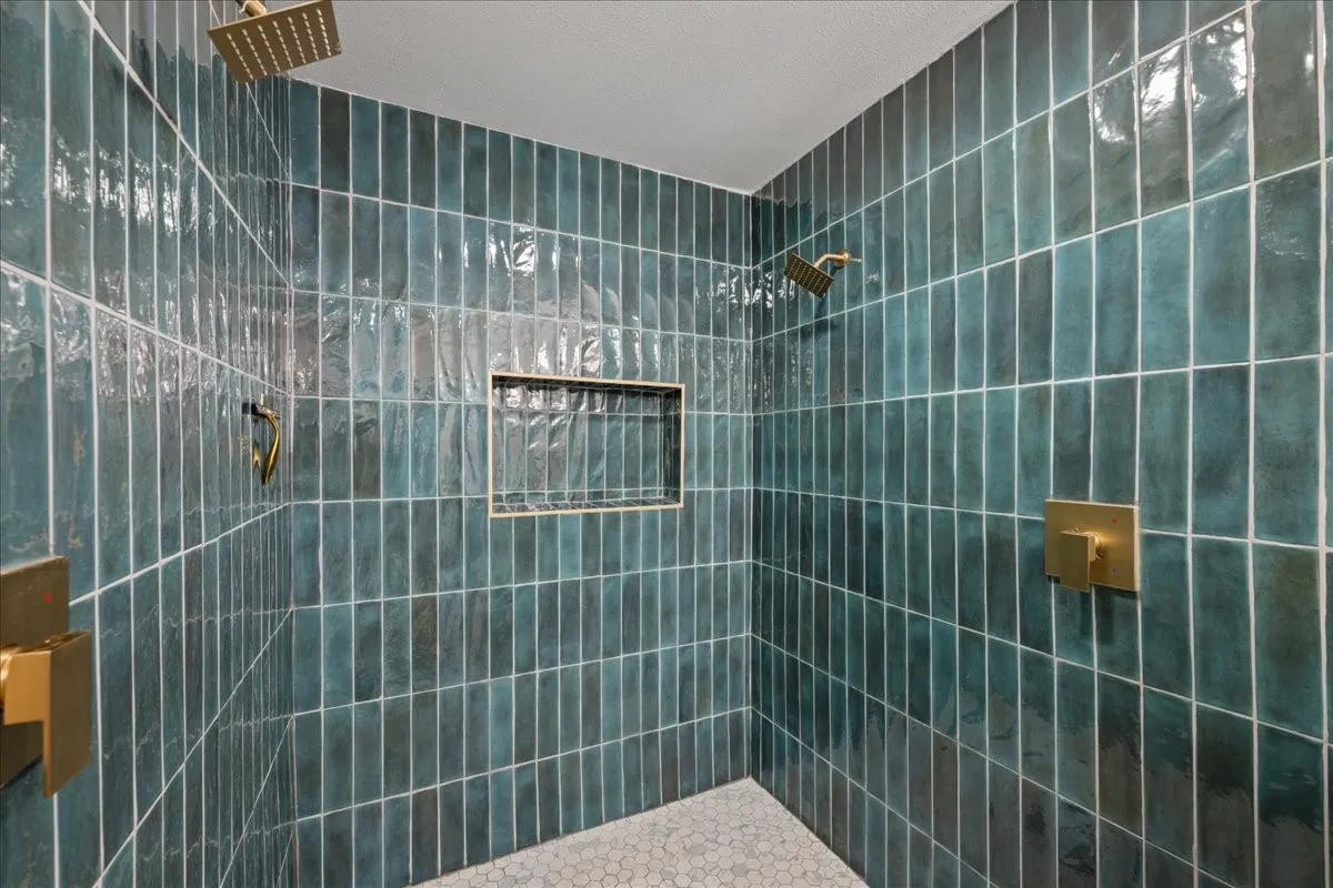Shower enclosure with vertical teal tile, dual rain shower heads, built-in recessed shelf, gold-finish fixtures, and mosaic tile flooring