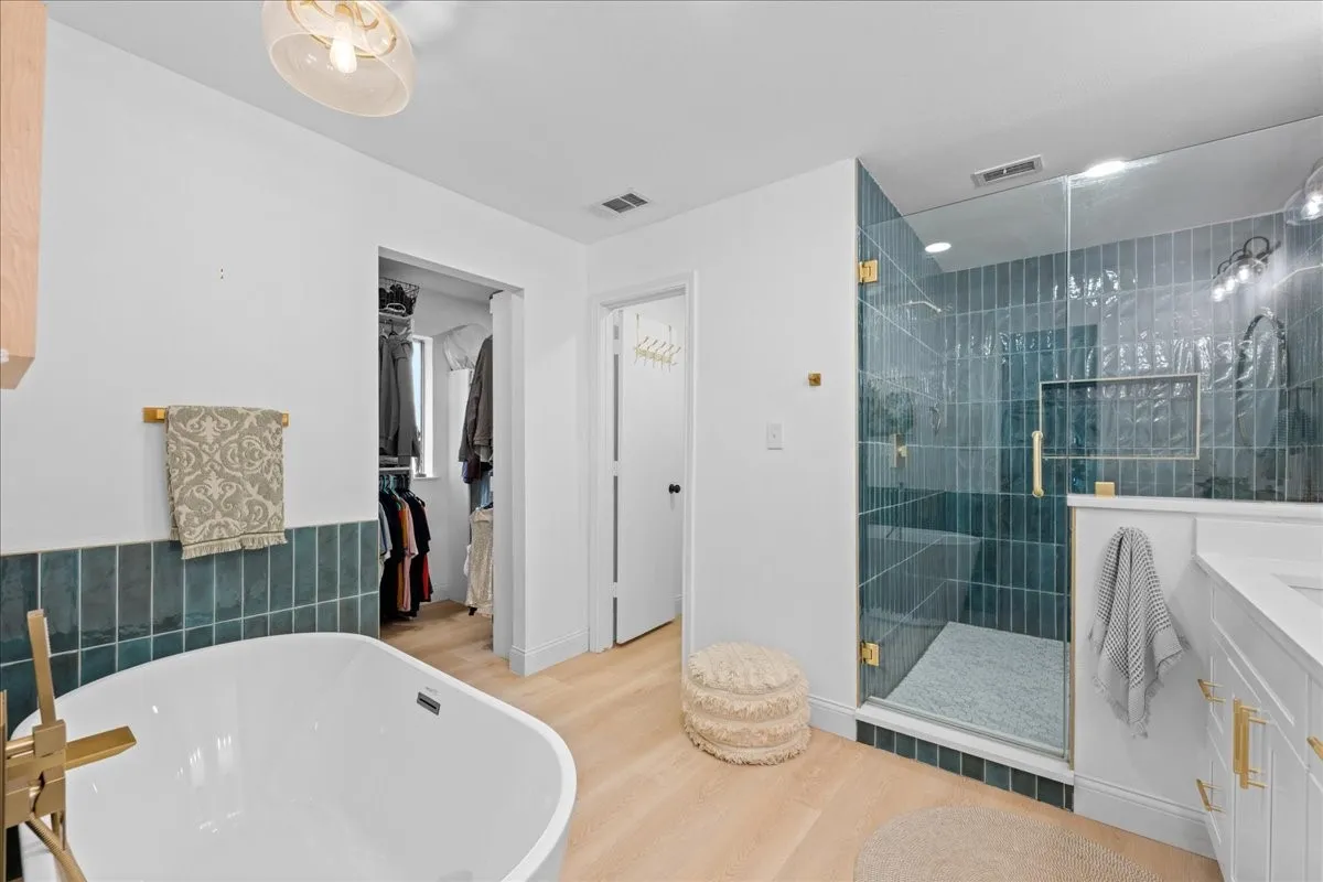 Freestanding soaking tub with brass fixtures, glass-enclosed shower with vertical teal tile, walk-in closet, wood-finish flooring, and a vanity with white cabinetry